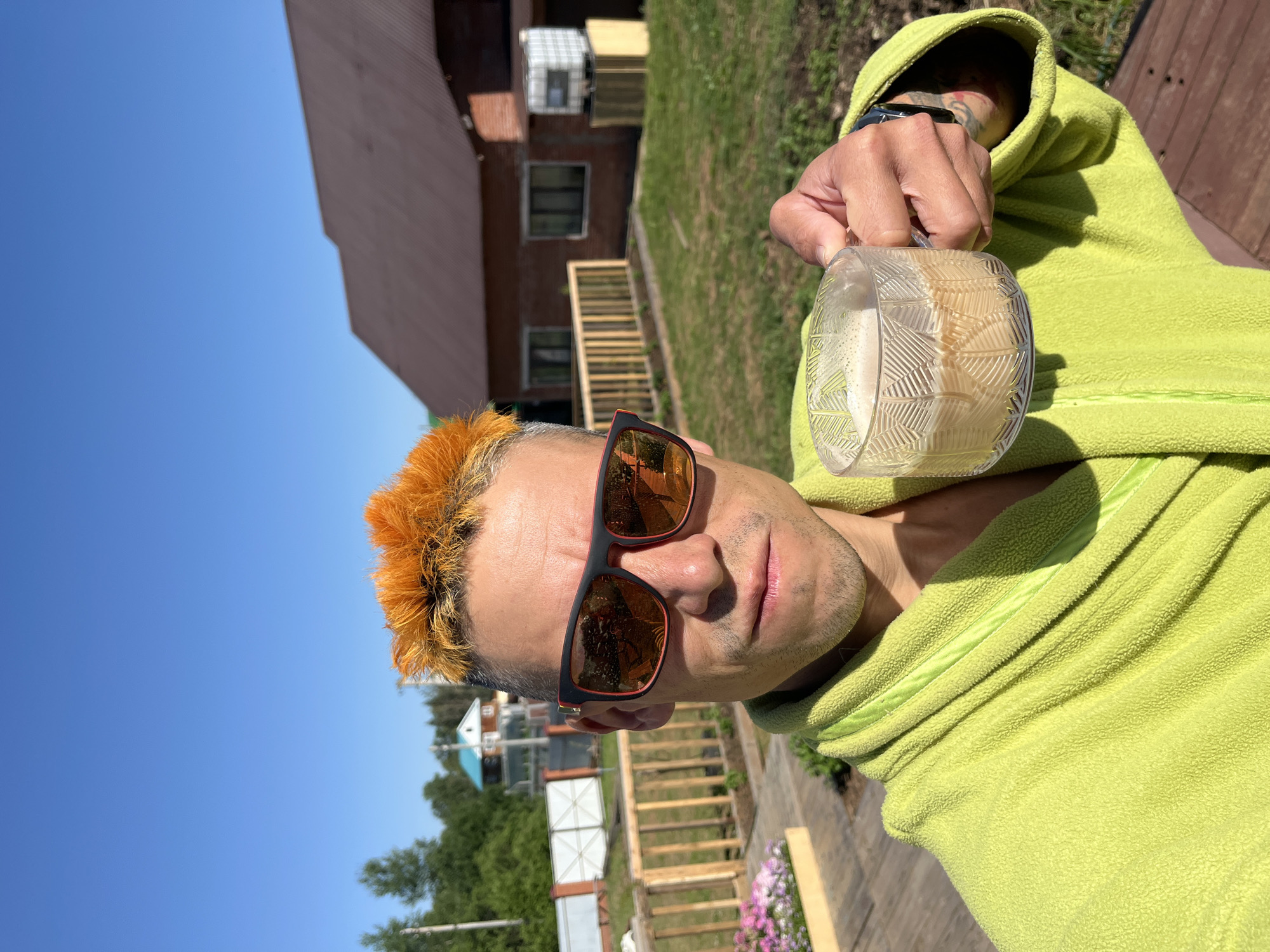 The image features a person with bright orange spiked hair and sunglasses, wearing a lime green hooded sweatshirt. They are holding a clear glass with a light-colored beverage, possibly a coffee or cocktail, in their right hand. The person is looking directly at the camera with a neutral expression.
The background suggests an outdoor setting with a clear blue sky. A rustic building with a red roof and several windows is visible, along with wooden stairs and some greenery. The overall impression is a casual, daytime scene, likely in a suburban or rural area. The image is captured from a slightly low angle, emphasizing the sky and the person's face.
There are no visible signs of text in the image. The lighting indicates it is a sunny day, and the shadows suggest the sun is high in the sky, pointing to midday or early afternoon. The setting appears to be in Chaikovsky, Russia, as indicated by the context provided.