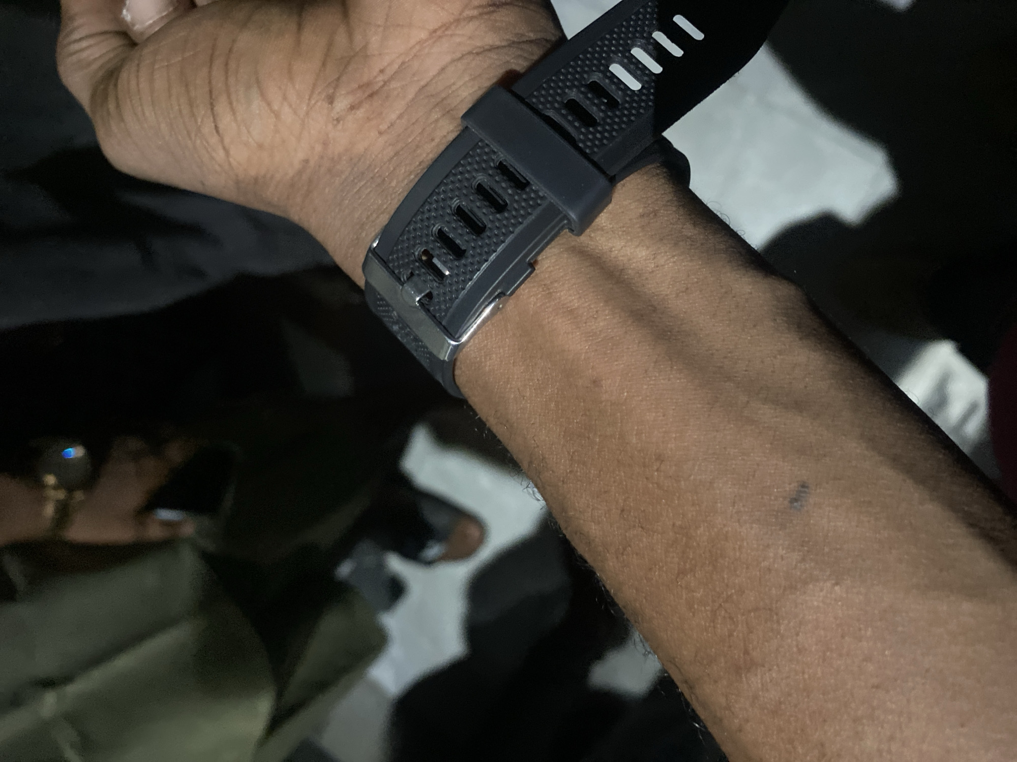 A close-up, eye-level shot captures a person's forearm, adorned with a dark gray, textured wristband watch. The watch band features a pattern of small raised dots and rectangular cutouts, secured by a metallic buckle and a dark gray loop. The skin of the forearm is brown, with visible pores and a scattering of fine hairs. A small, dark mark, possibly a smudge or a tiny mole, is visible on the skin. The background is dark and out of focus, with hints of lighter areas and shadows that suggest an indoor environment. The overall lighting is dim, with the watchband and forearm being the primary illuminated subjects. The image provides no information about the location in Sabon Gari West, Nigeria, or any specific actions or interactions occurring beyond the presence of the person and their watch.