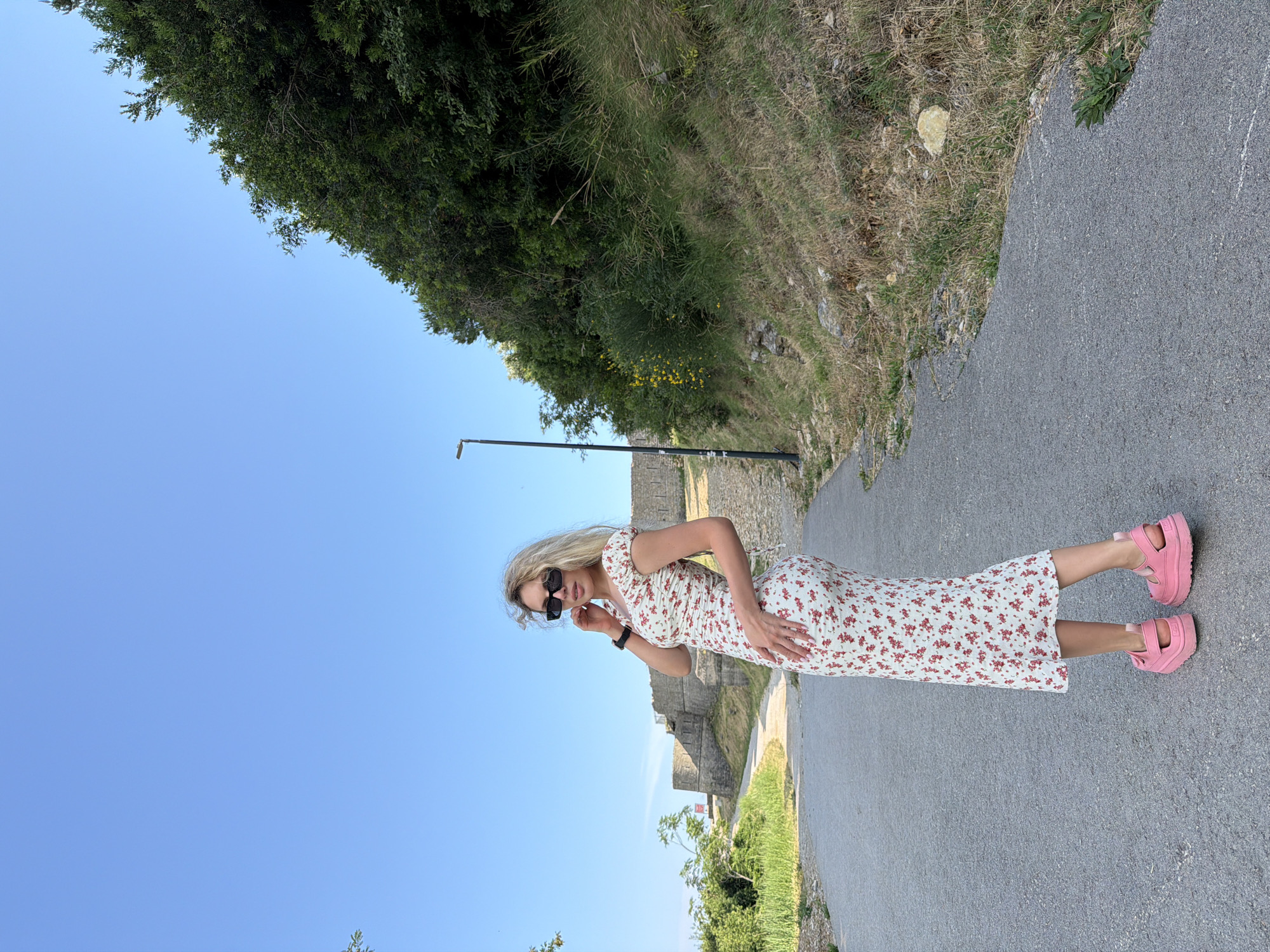 A woman with blonde hair and sunglasses stands on a paved path with a floral dress and pink platform sandals. She is positioned on the right side of the frame, facing slightly towards the camera. Behind her, a stone wall structure is visible on the left, with green foliage and trees surrounding it. The path curves to the left and ascends towards the stone structure. A tall black lamppost stands on the right side of the path, amidst the dense green vegetation that covers a sloping hill. The sky is clear blue. The location is identified as the Citadel Fortress in Budva, Montenegro.