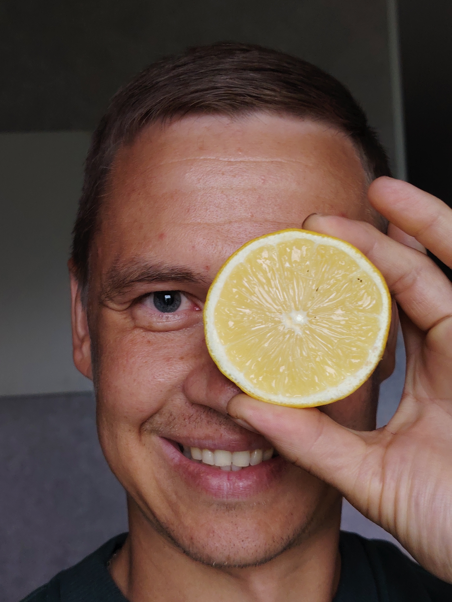 A close-up portrait features an adult man in an indoor setting against a plain, muted grey background. The man holds a bright yellow half-slice of a lemon playfully over his right eye, obscuring it from view. He is smiling broadly, revealing his white teeth, and looks directly at the camera with his visible blue left eye, conveying a happy and engaged expression.
The man has short, brown hair and a light-to-medium skin tone, with a slight stubble visible on his jawline and chin. The interior of the lemon slice is clearly visible, showcasing its segmented pulp. The lighting appears natural, suggesting it is daytime. There are no specific location cues visible within the image to indicate Chaikovsky, Russia, nor is any text present. The overall mood is lighthearted and cheerful.