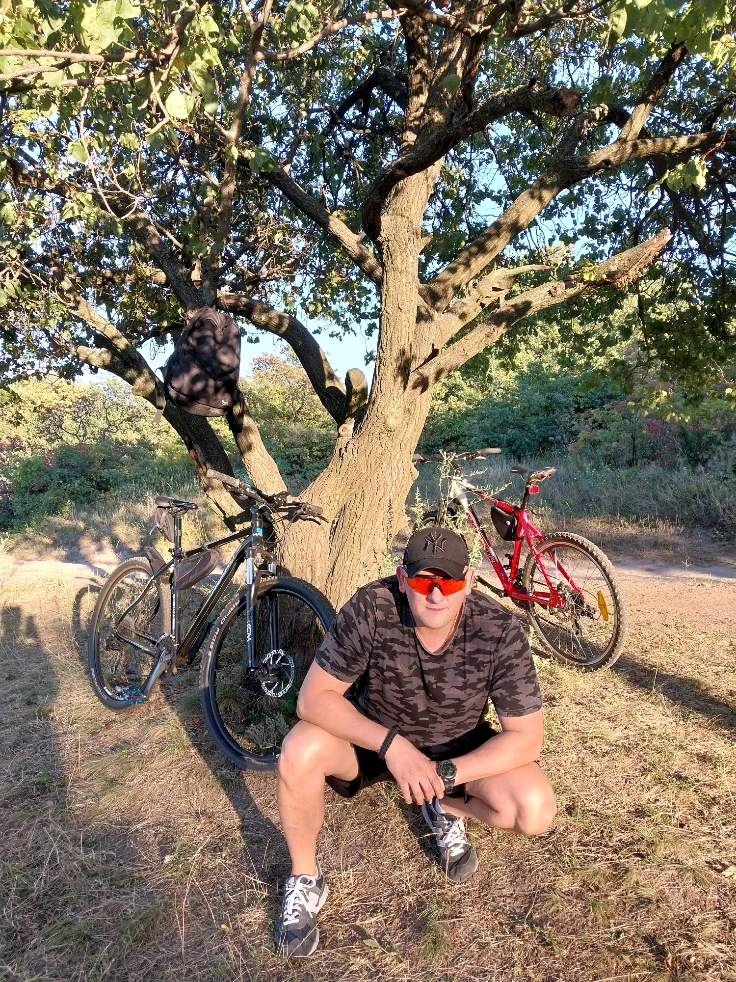In a sun-dappled outdoor setting, a man is crouched beside a large tree, two bicycles leaning against it. The man, wearing a camouflage t-shirt, black shorts, a black baseball cap, and orange-tinted sunglasses, is looking towards the camera. One bicycle, black and grey, is positioned to his left, its front wheel turned. Another bicycle, red, is leaning on the tree to his right. A black backpack is hanging from a branch of the tree above the man. The ground is covered with dry grass and dirt. The background shows dense foliage and trees. The location is identified as being in the city of Dnipro, Ukraine.