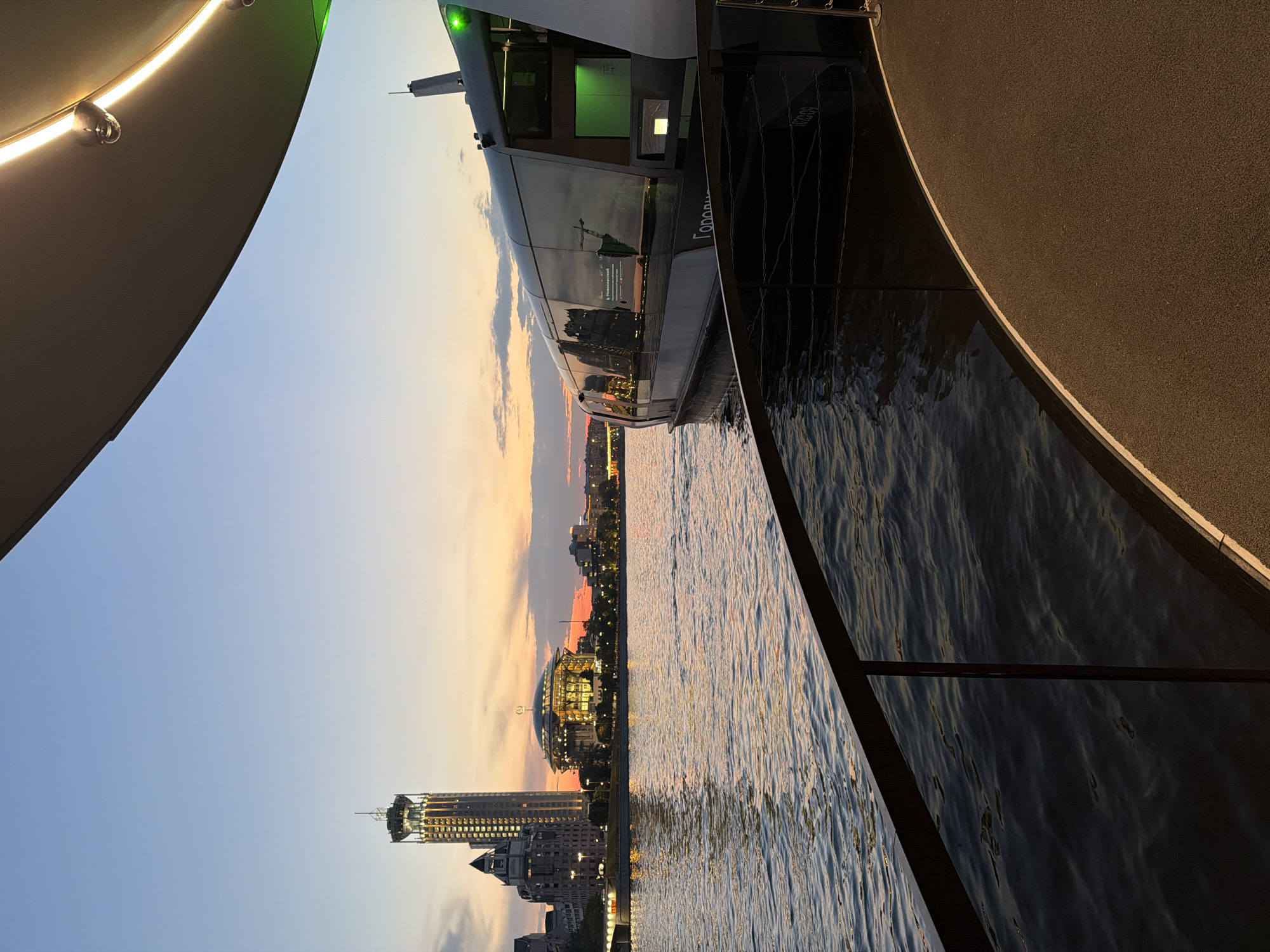 The image captures a serene sunset scene over the Moskva River in Moscow, Russia. In the foreground, a modern boat with a sleek, dark exterior and reflective windows is docked. The boat features illuminated signage and what appears to be green lighting. A dark railing separates the viewer from the water, and a textured pathway, possibly a deck, curves away to the right.
Across the river, the Moscow skyline is illuminated by the warm glow of the setting sun. Prominently featured are two distinctive buildings: a tall, slender skyscraper with a tiered top and illuminated windows, and a larger, domed structure that also appears to be lit from within. The water reflects the colors of the sky and the city lights, creating a shimmering effect. The sky transitions from a pale blue at the top to hues of orange and pink near the horizon, with scattered clouds adding texture. The overall atmosphere is tranquil and picturesque, suggesting an evening view from a river cruise or waterfront vantage point.