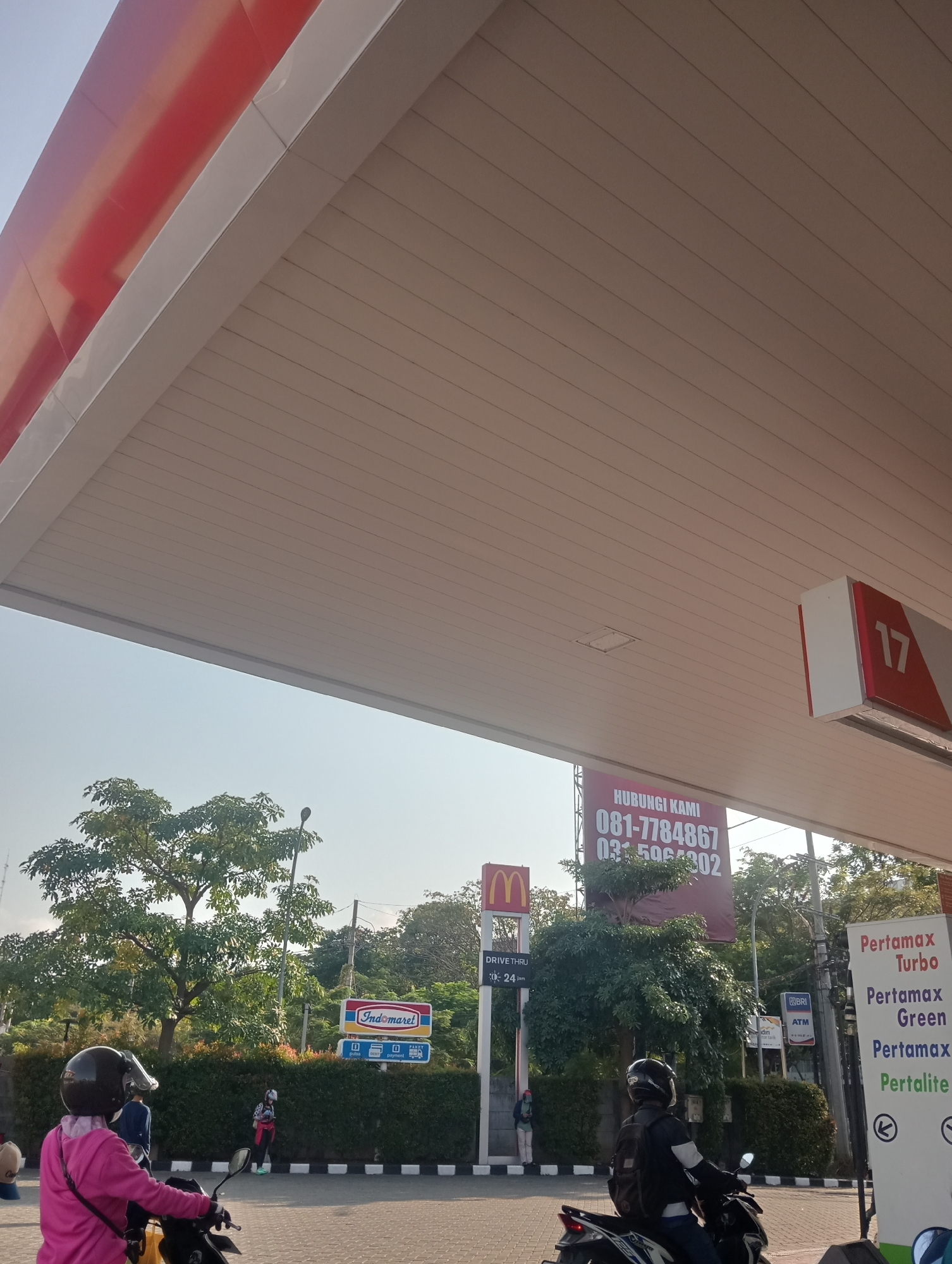 This image depicts a scene at a gas station in Surabaya, Indonesia, under a large white canopy. The canopy's underside is lined with beige panels, and its edges are accented with a red stripe. On the right side of the canopy, a red and white sign displays the number "17". Below the canopy, a large maroon billboard announces "HUBUNGI KAMI" (Contact Us) with phone numbers "081-7784867" and "031-5964302". To the left of this billboard, a McDonald's sign with the golden arches is visible, along with a "DRIVE THRU" sign indicating it is open "24 jam" (24 hours).
In the foreground, two individuals on motorcycles are present. On the left, a person wearing a pink hooded jacket and a black helmet is seated on a black motorcycle. On the right, another person wearing a black jacket and a black helmet with a backpack is also seated on a motorcycle. Several trees and green hedges form the background, with an Indomaret convenience store sign visible among them. Further back, a BRI ATM sign can be seen. The sky is clear and blue, suggesting a sunny day. The ground appears to be paved.