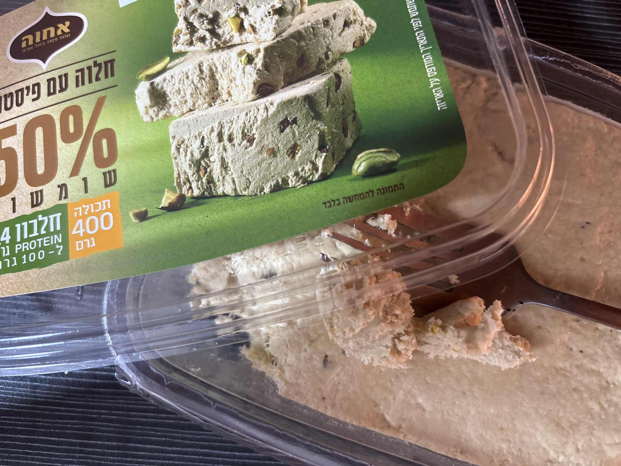 This is a close-up shot of a partially eaten container of halva with pistachios. The product is from the brand "Achva," established in Tel Aviv in 1929. The product packaging highlights "50% sesame" and "4g protein per 100g." A fork is used to take a piece of the halva from the container. The image is set on a dark-colored surface with a striped pattern. There is no indication this image was taken in บ้านบางรักษ์, Thailand.