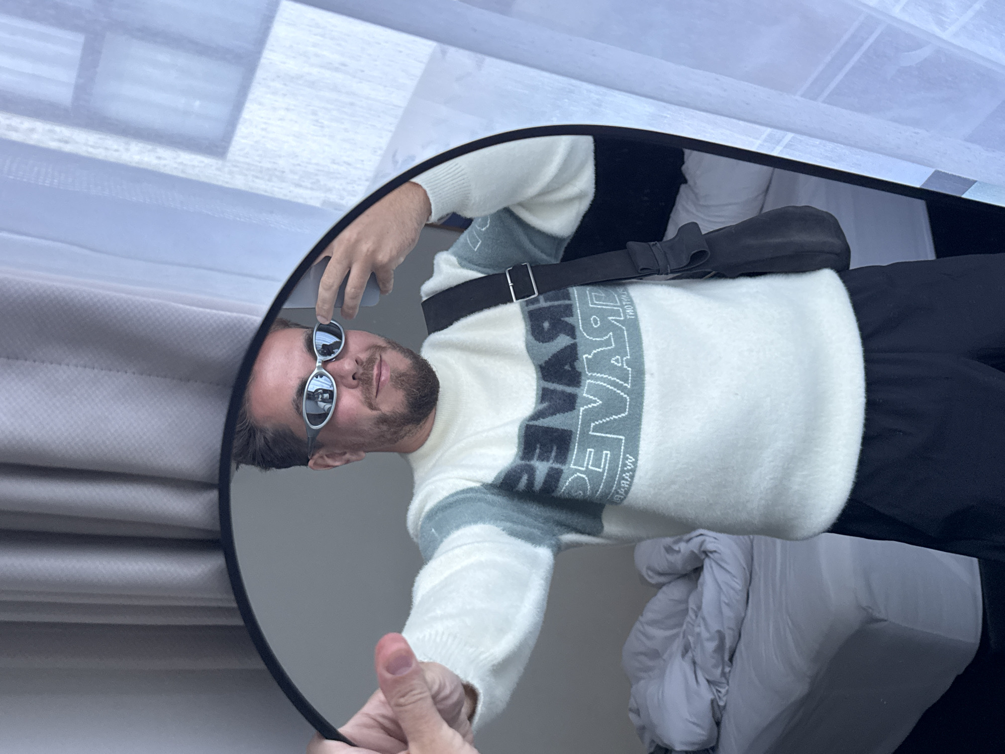 A man is reflected in a round mirror. The man is wearing sunglasses, a white sweater with a grey and black design, and black pants. He is also wearing a black crossbody bag. The environment around the mirror is a room with a bed and a window. The image does not provide information indicating its location is in North Kuta, Indonesia.