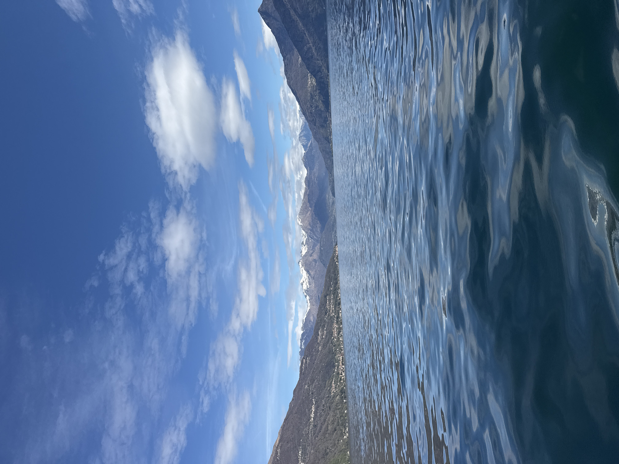 The image captures a view of Lake Como from Nobiallo, Italy. The scene is dominated by water with rippling surface, reflecting the sky. The distant shoreline reveals mountainous terrain with patches of snow at higher altitudes. The sky is a vibrant blue, dotted with scattered cumulus clouds.