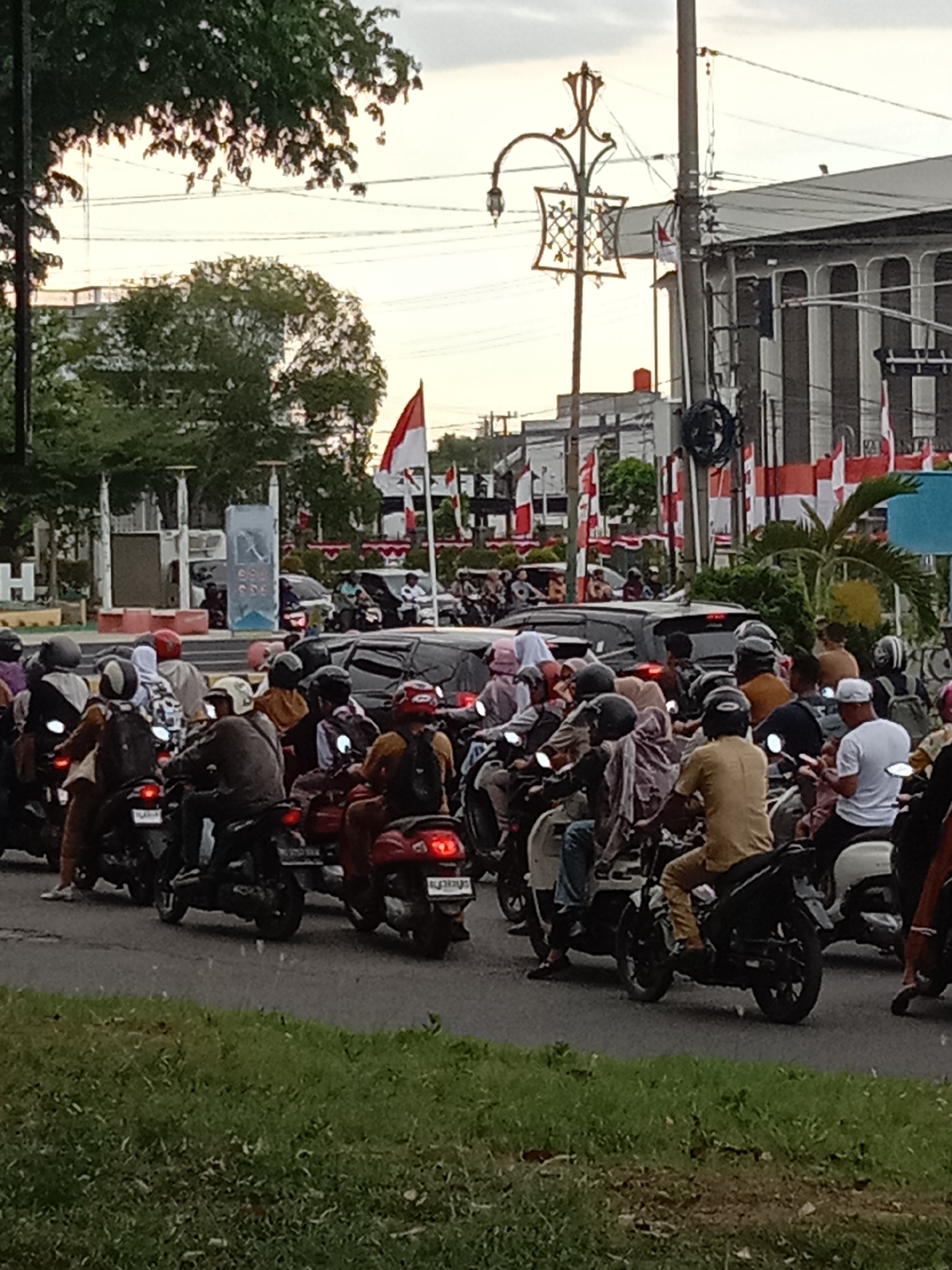 The image depicts a street scene in Lhokseumawe, Indonesia, characterized by heavy motorcycle traffic. In the foreground and midground, numerous motorcycles and their riders are present, forming a congested roadway. The riders are diverse in attire, with many wearing helmets and casual clothing, indicating typical urban commuting. Some motorcycles are red, while others are black and white. Several people are also visible as passengers on the motorcycles, often wearing headscarves or helmets.
In the background, larger vehicles, including cars, are part of the traffic flow. Beyond the vehicles, there are buildings with distinctive architecture, featuring vertical lines and arched windows, suggesting institutional or commercial structures. The presence of numerous Indonesian flags, both large and small, displayed on poles and attached to buildings, indicates a festive or patriotic occasion. The environment includes trees with lush green foliage and a cloudy sky, typical of tropical weather. A decorative lamppost with an ornate design is also visible on the left side of the image. The overall impression is one of a bustling urban environment during daylight hours, likely a public holiday or a significant event.