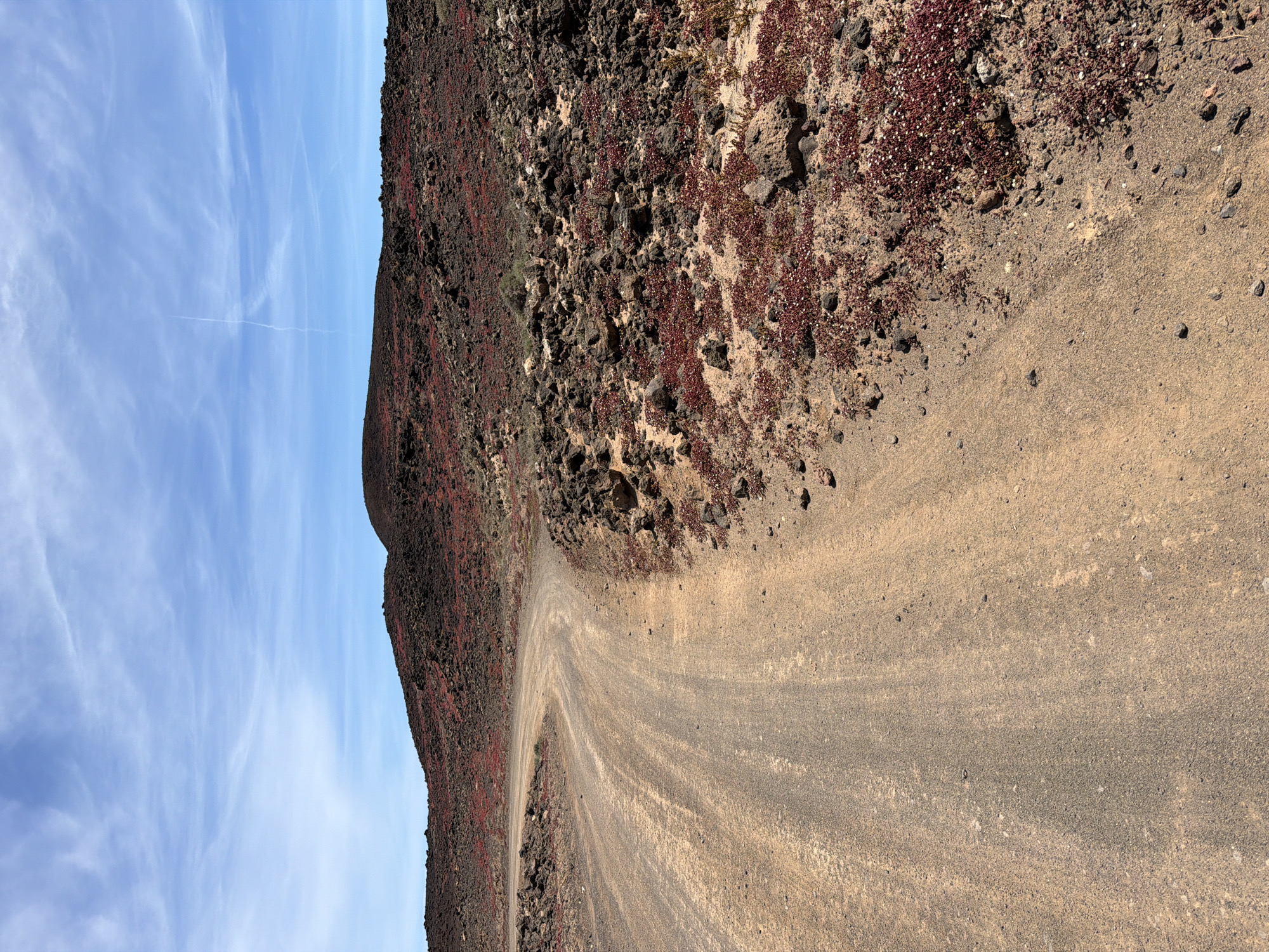 A dirt road curves around the base of a dark, rocky hill. The hill is covered with sparse red vegetation. The sky is blue with wispy clouds. This is possibly in Tinajo, Spain. No people are visible.
