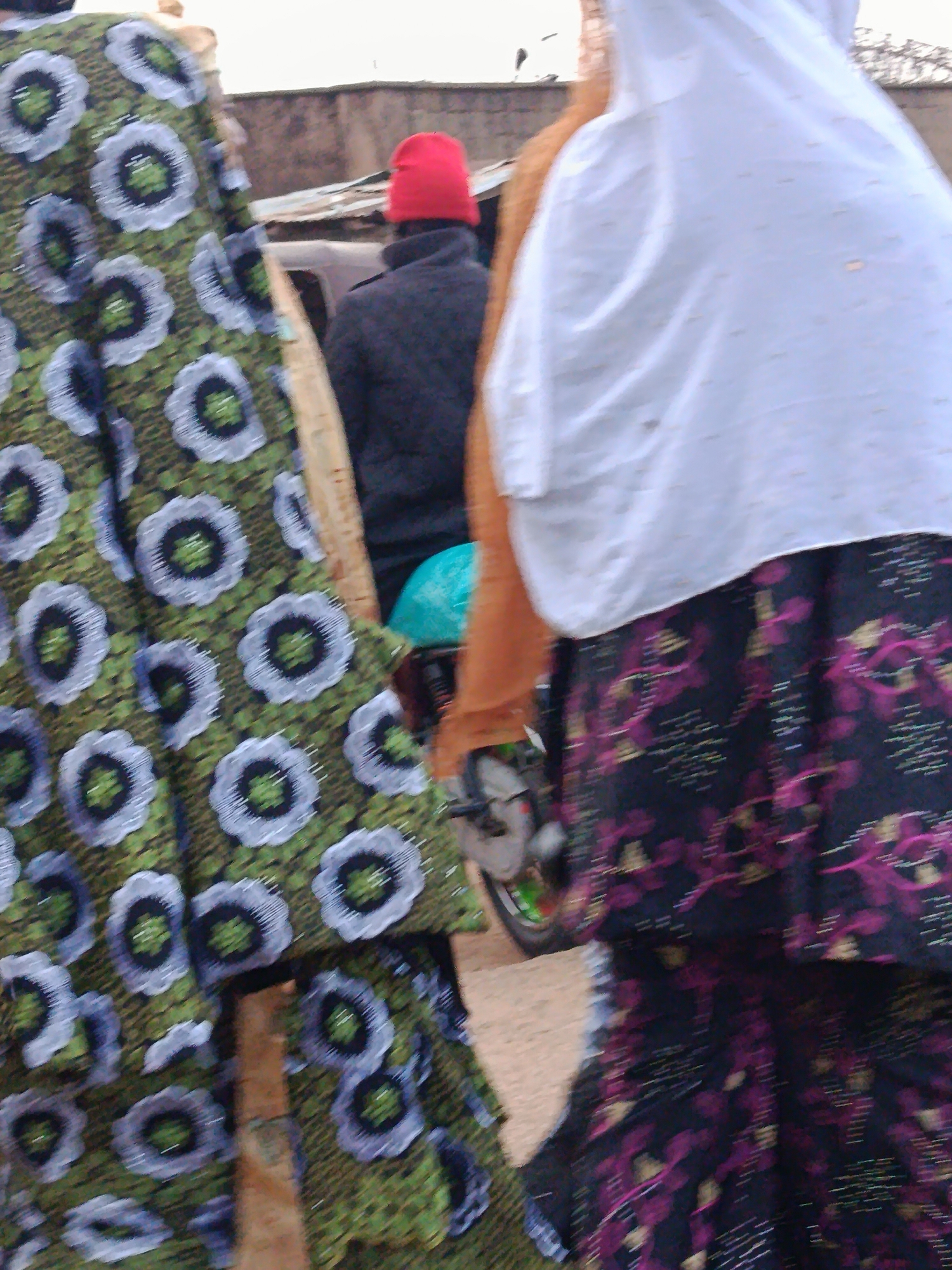 In New Stadium, Nigeria, a blurry image shows a man wearing a red beanie and a dark jacket, facing away from the camera. He is standing near a motorcycle. To the left of the man, a woman in a white hijab and a patterned green dress is visible, her body partially obscured by the dress. To the right, another woman, also partially visible, wears a dark skirt with a floral pattern and a white hijab. The background consists of a building and an overcast sky, suggesting an outdoor setting.