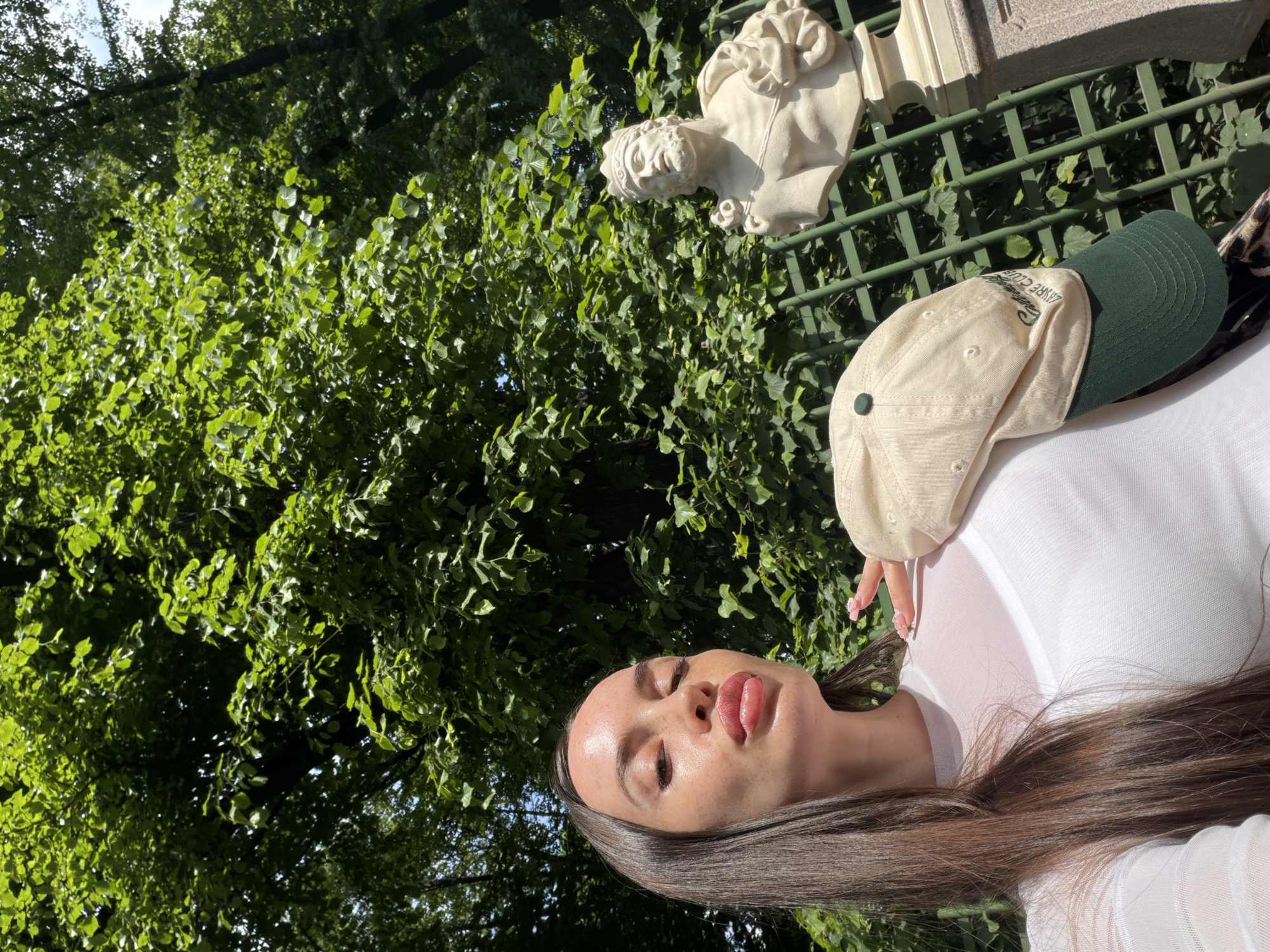 A young woman with long dark hair and fair skin is taking a selfie in a park. She is wearing a white mesh top with a white bra underneath and a beige baseball cap with a green brim and embroidery. She is puckering her lips and her left hand is resting on the cap. Behind her, a stone bust of a man wearing a crown stands on a pedestal. The bust is partially obscured by a green metal fence. Lush green trees and foliage form the background, suggesting a sunny day in a garden or park. The overall scene implies a casual outdoor setting, possibly within the grounds of a historical estate or museum in Saint Petersburg, Russia, such as the park of the Pavlovsk Palace or the grounds of the Peterhof Palace, known for their sculptures and gardens.