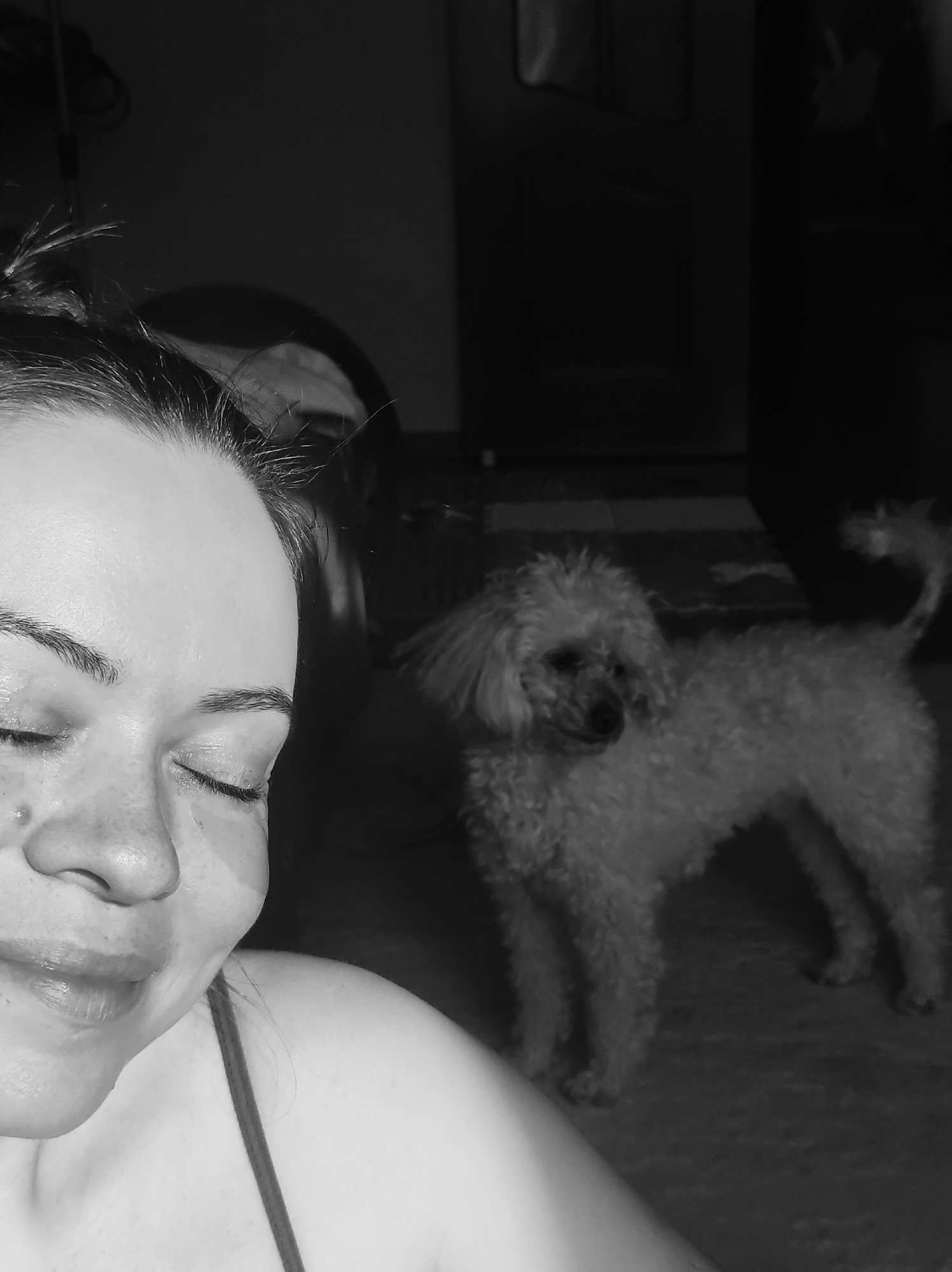 A black and white image captures a close-up of a woman with a small, fluffy dog in the background, set indoors in Chaikovsky, Russia. The woman occupies the left side of the frame, with her eyes gently closed and a soft, peaceful expression, hinting at a slight smile. Her face is well-lit, showing details of her features and some wisps of hair pulled back from her forehead. She is wearing a top with a thin strap visible on her shoulder.
To the right, in the mid-ground, a light-colored, curly-haired dog, resembling a poodle, stands. The dog is looking slightly away from the camera, its head turned to the right. Its body appears to be facing more towards the viewer. The background is dark and indistinct, suggesting a dimly lit indoor space, possibly a home, and implying the time of day could be evening or night. The woman is in sharp focus, while the dog and background are softer, indicating a shallow depth of field. The scene conveys a quiet, intimate moment between the woman and her pet. No visible text is present in the image.