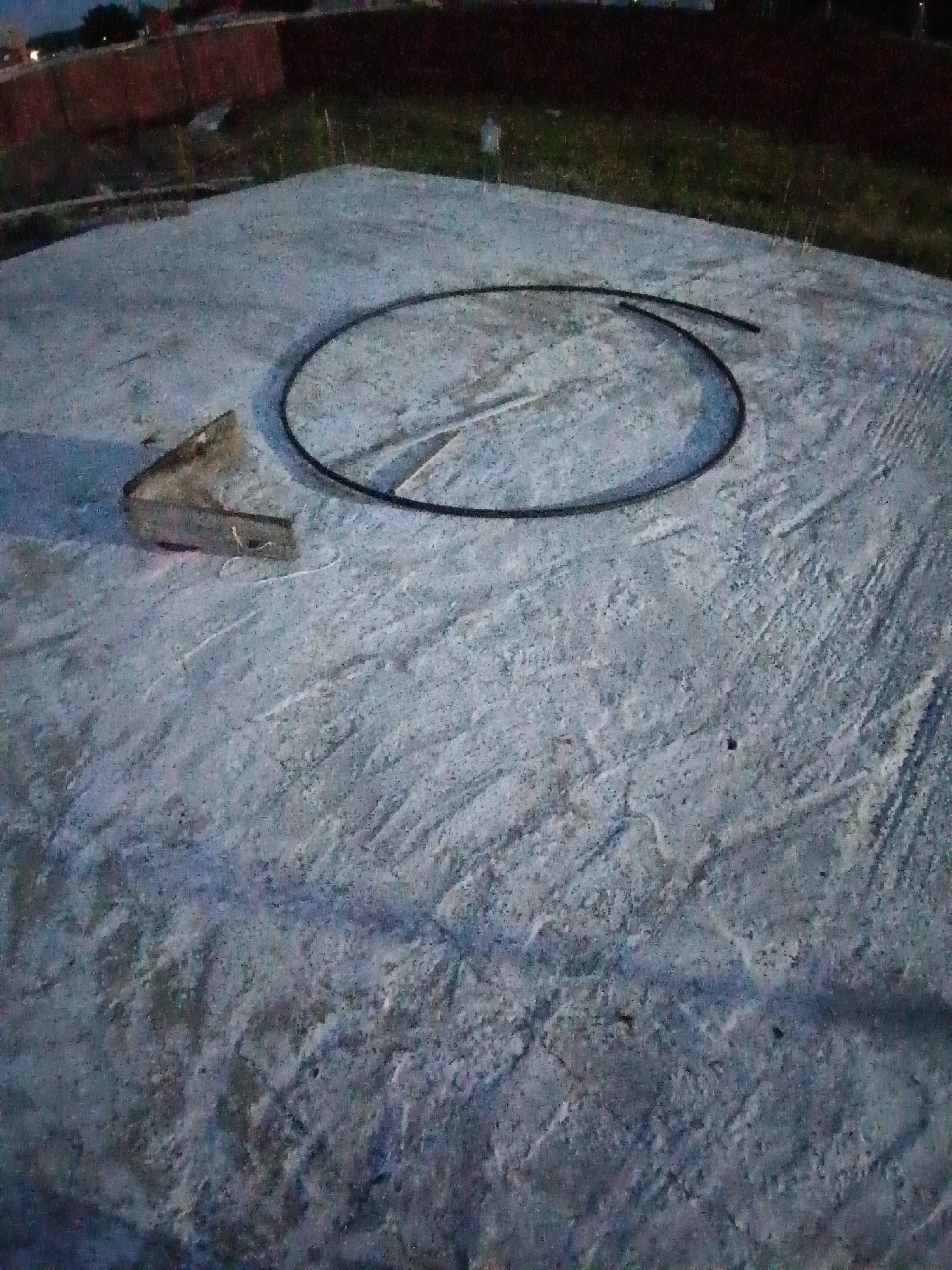 A circular black hose lies on a rough, grey concrete surface, forming an incomplete circle. To the left of the hose, a small, metallic object with a triangular opening is placed on the concrete. The concrete surface appears textured and uneven. In the background, a dark embankment with sparse green vegetation is visible, and beyond that, blurred structures and lights suggest an urban environment. The lighting in the image is dim, indicating it might be dusk or nighttime. The scene appears to be outdoors. The location is specified as the city of Amursk, Russia.