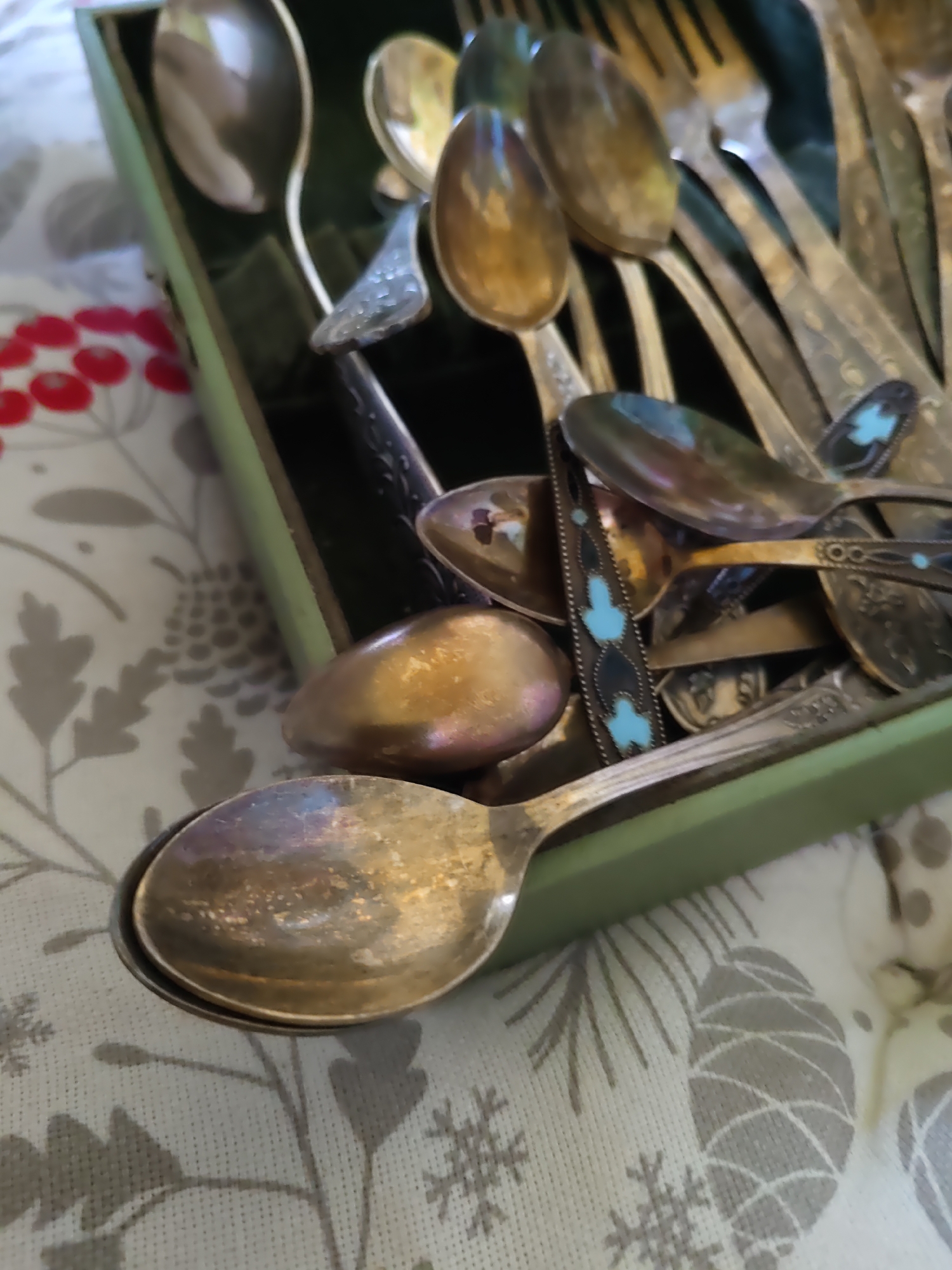 The image shows a collection of antique silver spoons and forks resting in a green velvet-lined box. The cutlery is tarnished and some pieces have intricate designs, including one spoon with inlaid turquoise. The box is placed on a tablecloth with a festive, possibly Christmas-themed, pattern of pinecones and snowflakes. The lighting is dim, creating reflections on the metal surfaces. There are no people visible in the image. The scene appears to be an indoor setting, possibly a dining room or display cabinet, in Rostov-on-Don, Russia.