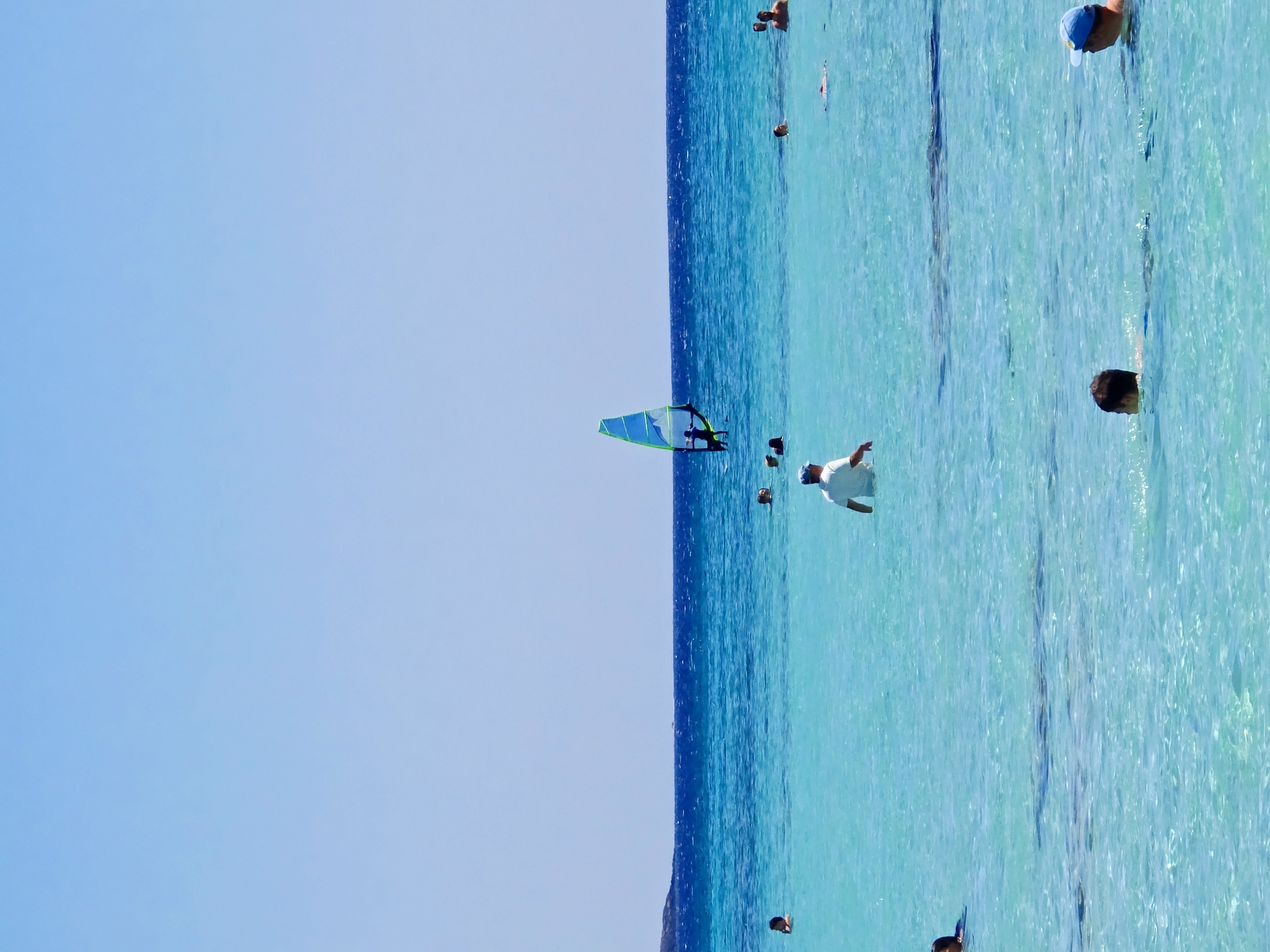 The image depicts a clear, sunny day at a beach with turquoise water. In the distance, a windsurfer is seen with a blue and green sail. Several people are scattered throughout the water, some swimming and others standing. The water is shallow in the foreground, with visible ripples and the sandy bottom beneath. The sky is a bright, cloudless blue. There is a landmass in the far distance on the left. The overall scene is tranquil and recreational.