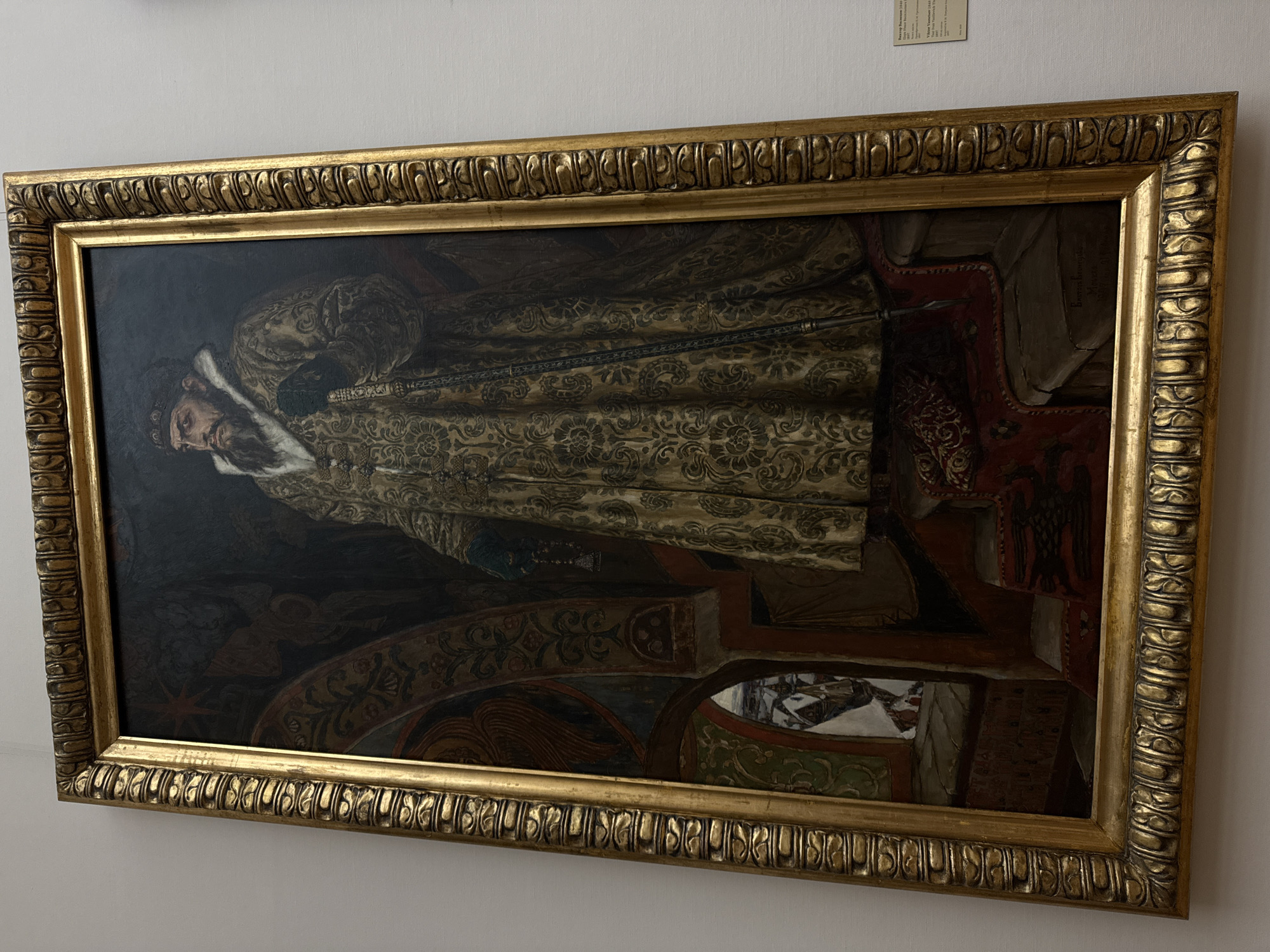 The image shows a framed oil painting of a standing, bearded man in regal attire, possibly a tsar. He wears a heavily patterned gold robe with fur trim. The man is holding a staff and standing on a red-draped platform with decorative elements. The background is dark and ornate. The painting is displayed on a white wall inside a building that appears to be a museum, possibly located in Moscow, Russia, given the style of the art.