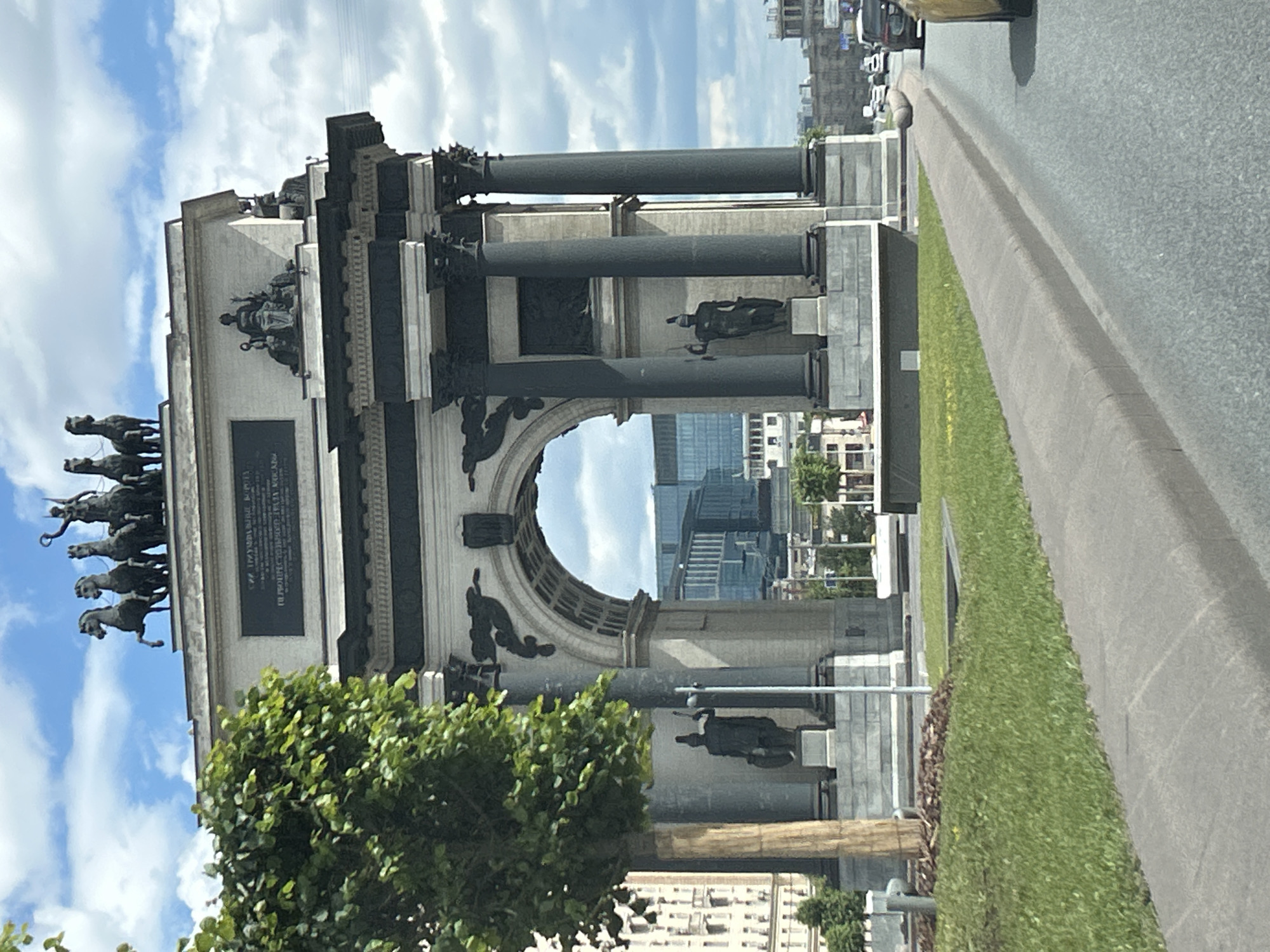 The image shows the Triumphal Arch in Moscow, Russia, viewed from a street level perspective, likely from a moving vehicle. The arch is a grand structure with classical architectural elements, including columns, an archway, and sculptures. Atop the arch, a quadriga (a chariot drawn by four horses) is prominently displayed, with a figure driving it. Below the quadriga, there's an inscription on a plaque. Two large, dark columns flank the archway, and on either side, at the base of the arch, stand statues of soldiers holding shields and spears.
The environment is urban, with a tree on the left side partially obscuring the view of the arch. In the background, modern buildings with glass facades are visible through the archway, suggesting a mix of historical and contemporary architecture. The sky is partly cloudy with white clouds against a blue background. The foreground shows a paved road with a grey curb and a patch of green grass. Cars are visible on the road in the distance, indicating it is a public street. The lighting suggests it is daytime.