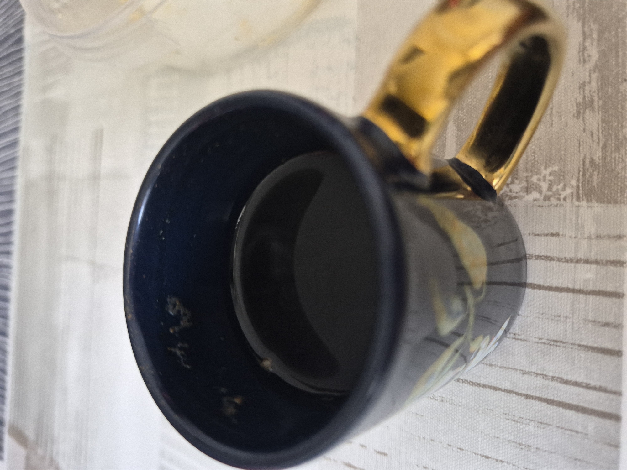 A close-up, eye-level shot shows a dark blue mug with a gold handle filled with black liquid, likely coffee. The mug is placed on a light-colored, textured surface, possibly a placemat or tablecloth. Some debris is visible on the inside rim of the mug. To the right of the mug, a portion of a clear plastic container with a light-colored filling is visible. The background is out of focus, with horizontal lines suggesting a surface or blinds. No people or animals are present in the scene. The lighting appears to be from overhead.
