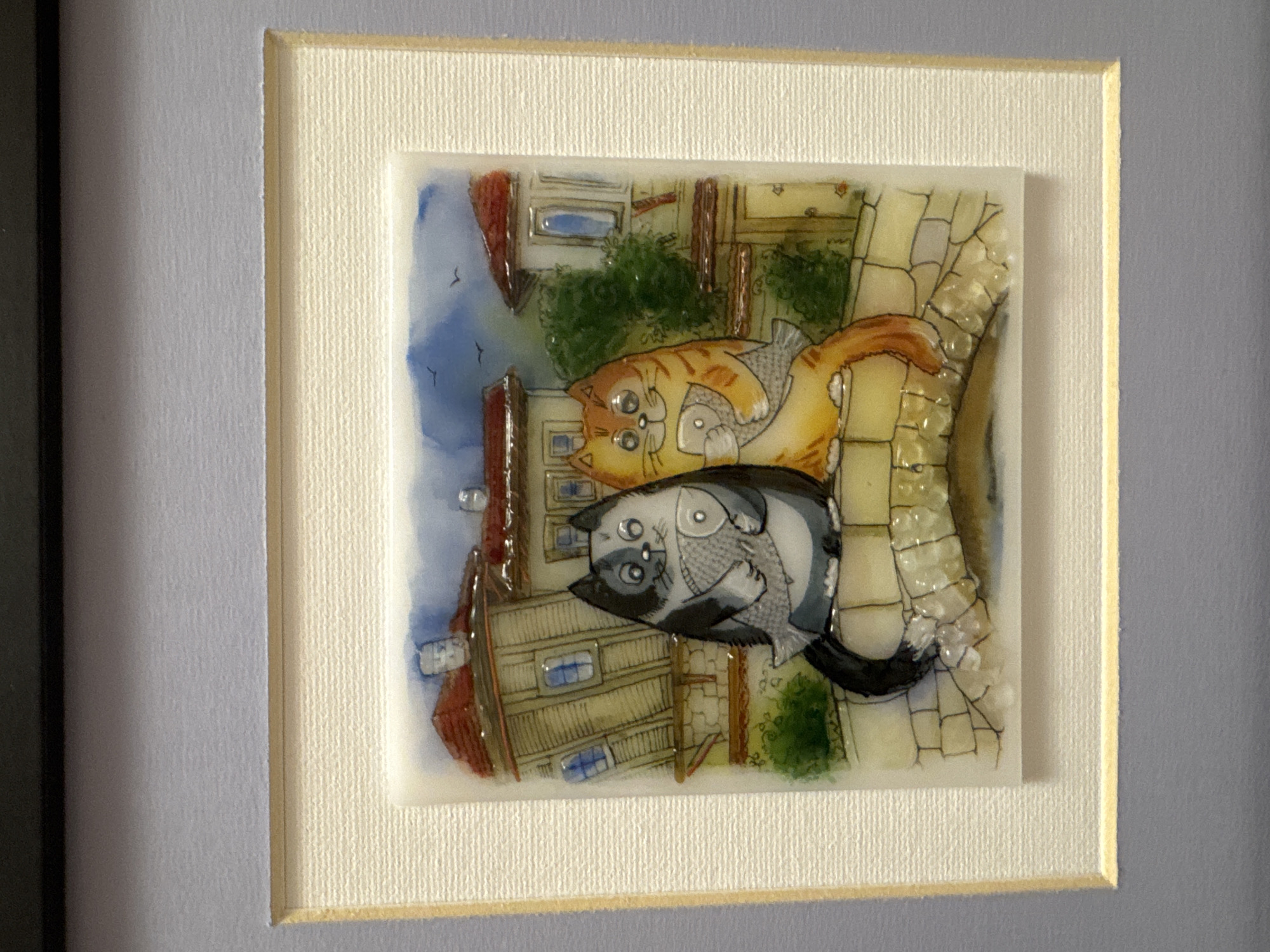 A framed art piece depicting two cats and a fish is presented. The upper cat is orange, and the lower one is gray and white, clutching a fish. The background suggests a cityscape, featuring buildings with red rooftops and blue windows. The art piece is mounted in a white frame with a textured surface, which is then set within a larger gray frame. The location, Plovdiv, Bulgaria, is inferred from the architectural style depicted in the art piece.