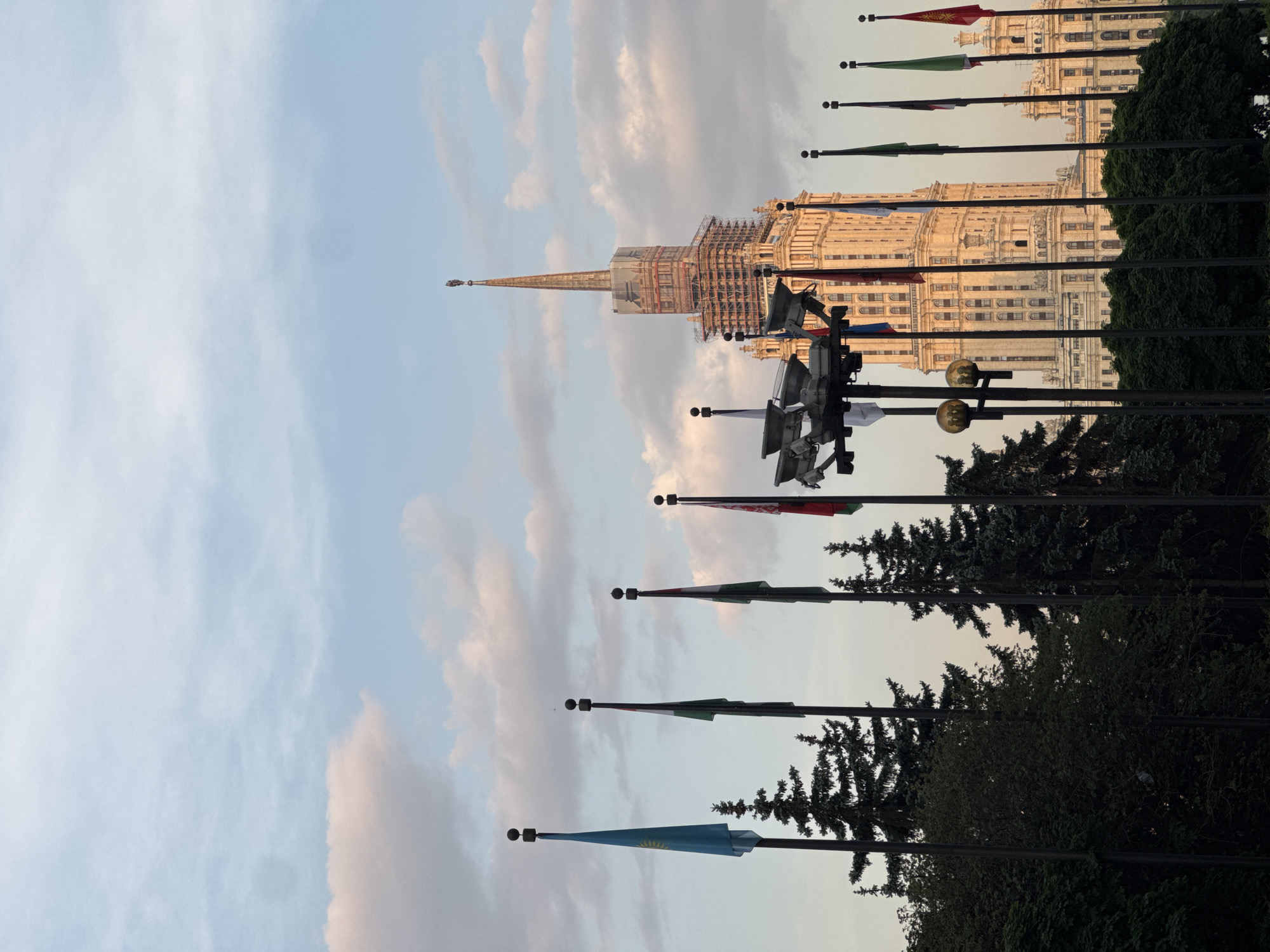 The image shows the Ministry of Foreign Affairs building, one of the Seven Sisters skyscrapers, in Moscow, Russia. The building is visible behind a row of flagpoles with various national flags. The sky is cloudy with pale blue and pink hues. The lower portion of the image is obscured by trees and bushes. The top of the building appears to be undergoing some form of construction or renovation, with scaffolding visible around the spire. There is a television camera located on one of the flagpoles.