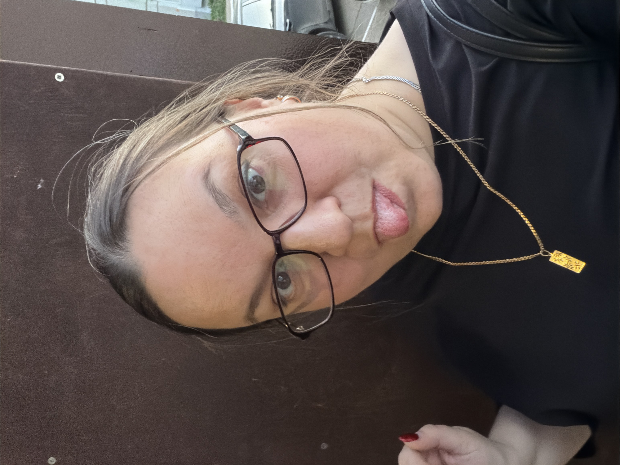 A woman wearing glasses is sticking her tongue out in a selfie. She has a gold necklace and a black shirt on. She is on what appears to be brown painted metal stairs. Based on the prompt, this image was taken in Chaikovsky, Russia.