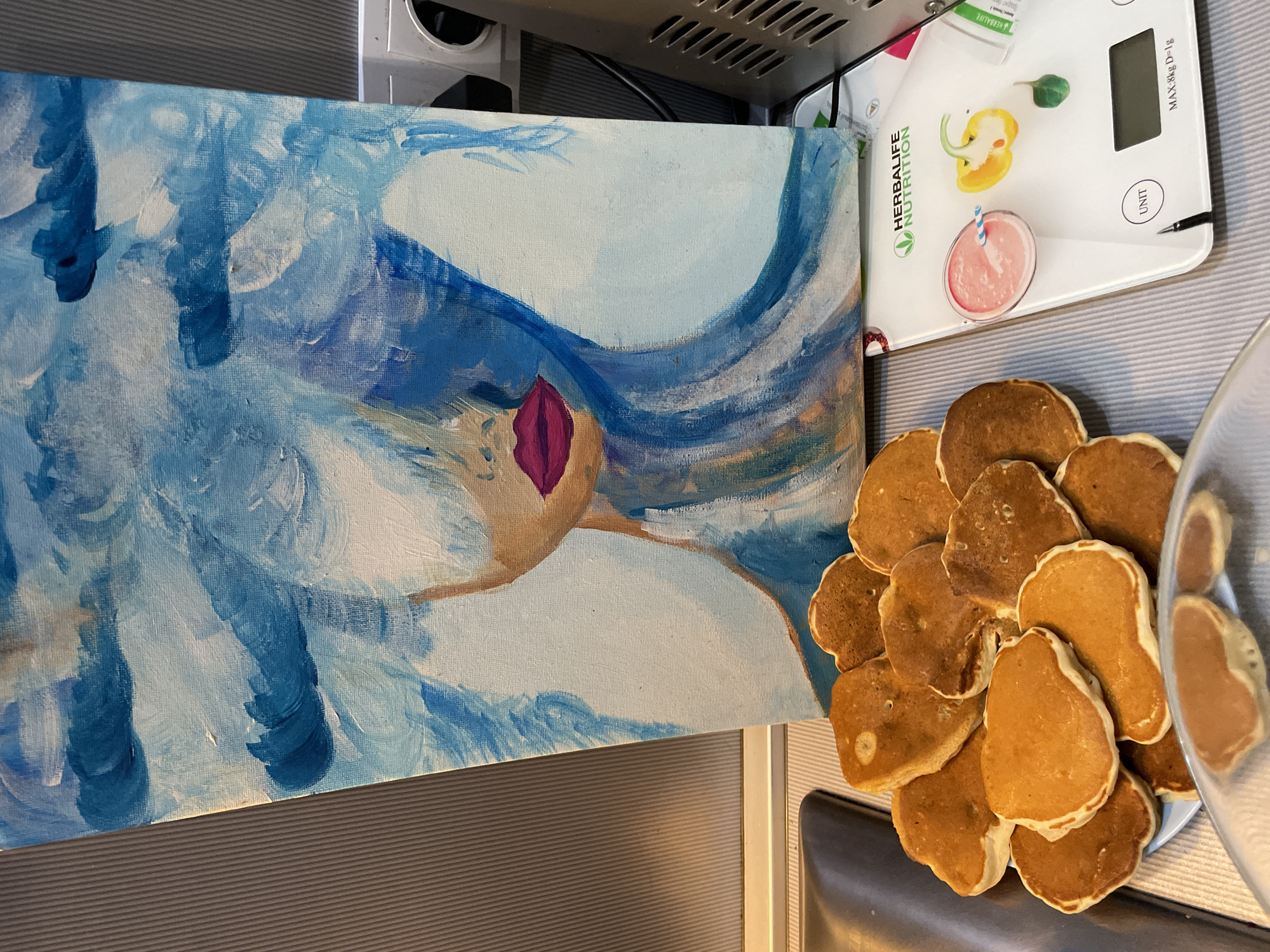 The image depicts an indoor scene in the city of Sochi, Russia, likely a kitchen or dining area. A stylized painting on canvas, primarily in shades of blue, white, and brown, occupies the central background. The painting features a partial depiction of a female face with exposed light brown skin around the chin and dark pink lips; the upper face and eyes are obscured by broad, flowing brushstrokes of blue and white, resembling hair or waves. Darker blue cloud-like shapes are present in the upper left of the artwork.
In the lower left foreground, a white circular plate holds a stack of approximately 15-20 small, golden-brown pancakes of varying irregular shapes. A few additional pancakes are visible on a reflective surface in the immediate foreground. To the lower right, a white digital kitchen scale branded "HERBALIFE NUTRITION" is positioned. Its display reads "MAX:8kg D=1g" and features a "UNIT" button. On the scale platform, a clear glass contains a pink liquid with a blue and white striped straw. Next to the glass, an artificial yellow bell pepper, cut in half, and a small artificial green leaf are present. A white container, partially obscured, is visible behind the scale.
The items rest on a light grey surface with horizontal ridges. A dark grey, smooth, curved appliance is partially visible on the far left. The background features a light grey wall with vertical indentations to the left of the painting. To the right, a white electrical outlet with a dark circular opening is visible, and below it, a metallic appliance with horizontal grilles is partially captured. No individuals are present in the scene.