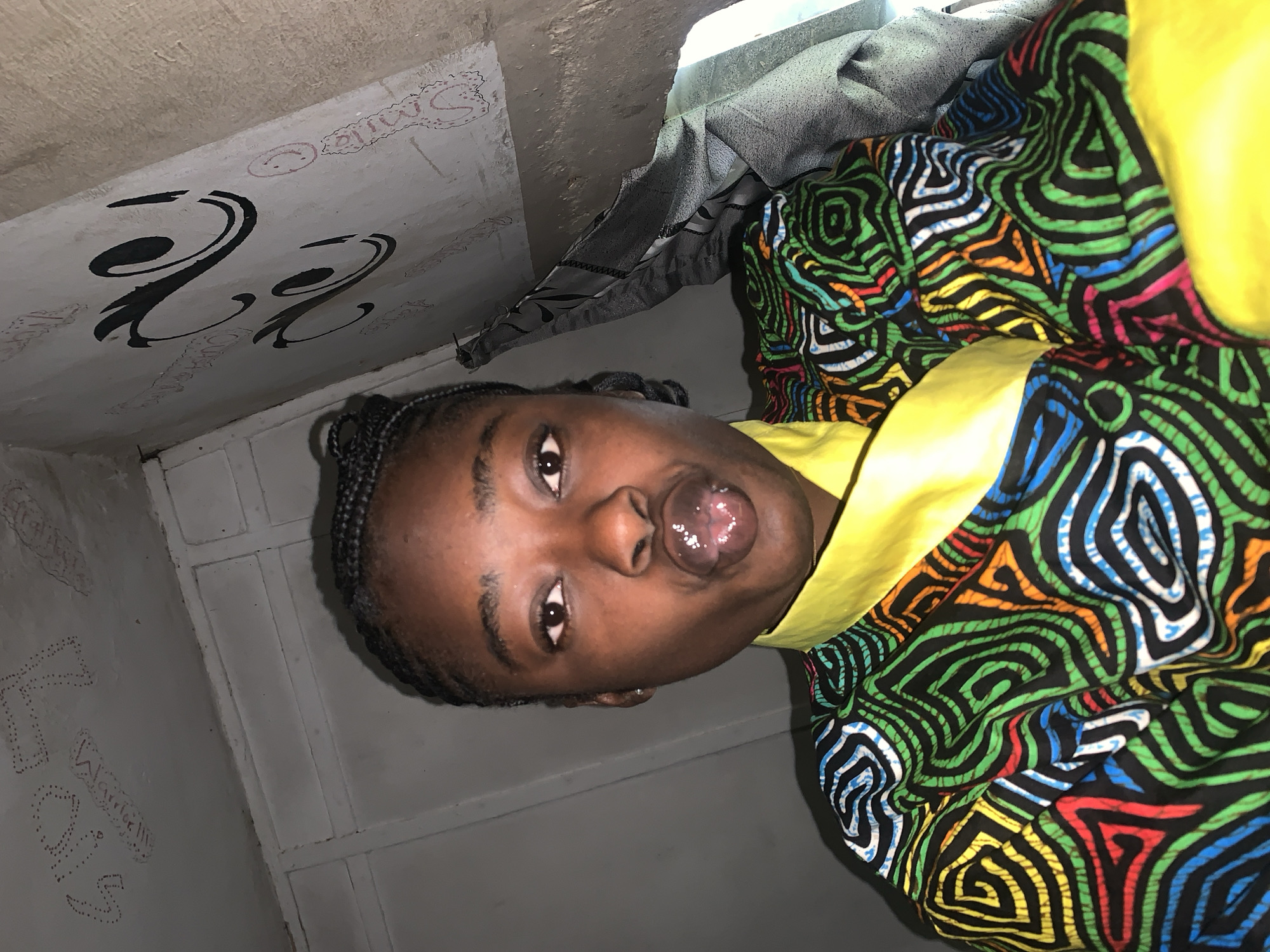 A young Black woman with braided hair is taking a selfie in what appears to be an indoor space. She is wearing a colorful patterned shirt with a yellow collar. Her lips are pursed and appear glossy. The background shows a textured wall with some faint markings and what looks like a piece of gray fabric draped to the right. The overall lighting is dim. The location is not ascertainable from the image.