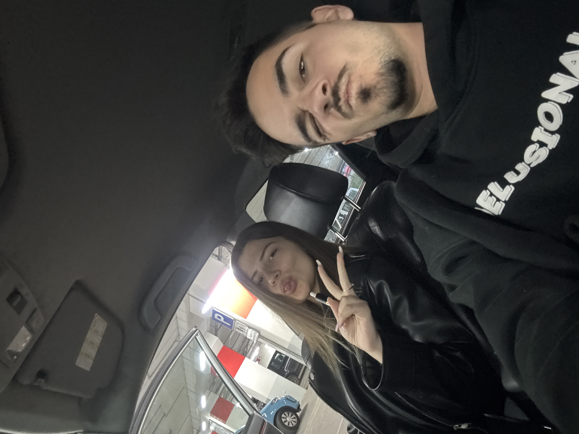 The image features a selfie of two individuals, a male and a female, inside a car that appears to be parked in an indoor or underground parking garage in Sofia, Bulgaria. The photograph is taken from a low, upward-angled perspective.
The male, positioned towards the top of the frame, has dark hair and a beard. He is wearing a black hoodie with the word "ELUSIONAL" partially visible in white text on the front. He looks towards the camera with a playful, puckered lip expression.
The female, seated in the passenger side below and to the right, has long, brown hair and is wearing a black leather jacket. She also has a puckered lip expression and holds up a peace sign with her left hand. Her nails are long and light-colored. She is holding a slim, dark, pen-like object, possibly a vape device. A ring is visible on her finger.
The car interior elements such as the ceiling, sun visor, and a seat headrest are visible. Through the car window, elements of the parking environment can be seen, including a light blue car, red and white striped support pillars, and artificial overhead lighting. A blue square sign with a white "P" clearly indicates a parking zone. The overall scene suggests a casual moment captured in a brightly lit parking facility.