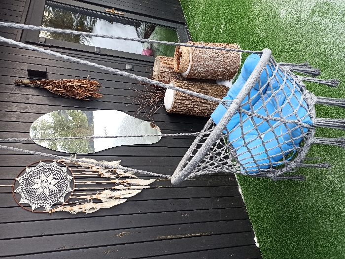 An outdoor scene in Nizhneye Zadolgoye, Russia, features a recreational area set against a dark, vertically paneled wooden wall. In the foreground, a grey macrame hanging chair with blue cushions is suspended, showing a light dusting of snow on its surface. The chair hangs over a surface covered with green artificial turf, which is also lightly and irregularly covered with snow. On the wooden wall, from left to right, are a large, circular dreamcatcher with intricate white crochet work and numerous white feathers; an irregular, organic-shaped mirror reflecting a snow-covered outdoor landscape with trees; and a bundle of dried organic material, possibly a venik, hanging from a hook. A black wall-mounted fixture is positioned to the right of the mirror. At the base of the wall, on the snow-dusted turf, three tree stumps of varying heights are positioned as decorative or functional elements, with some snow accumulation on their tops. A cluster of bare twigs is also visible near the stumps. A dark-framed glass door, reflecting more of the snowy exterior, is partially visible on the far right of the scene.