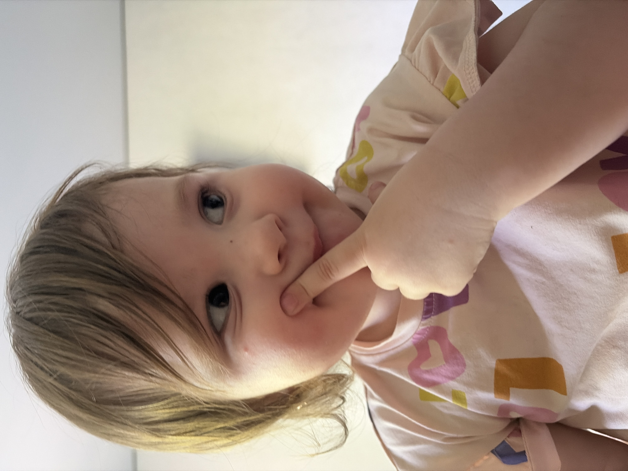 A toddler with blonde hair and fair skin is centered in the frame, angled to the left. She is wearing a light pink shirt with colorful, abstract patterns. The child's left index finger is touching her cheek. The background consists of a plain, light-colored wall. No details suggest the location is Chaikovsky, Russia.