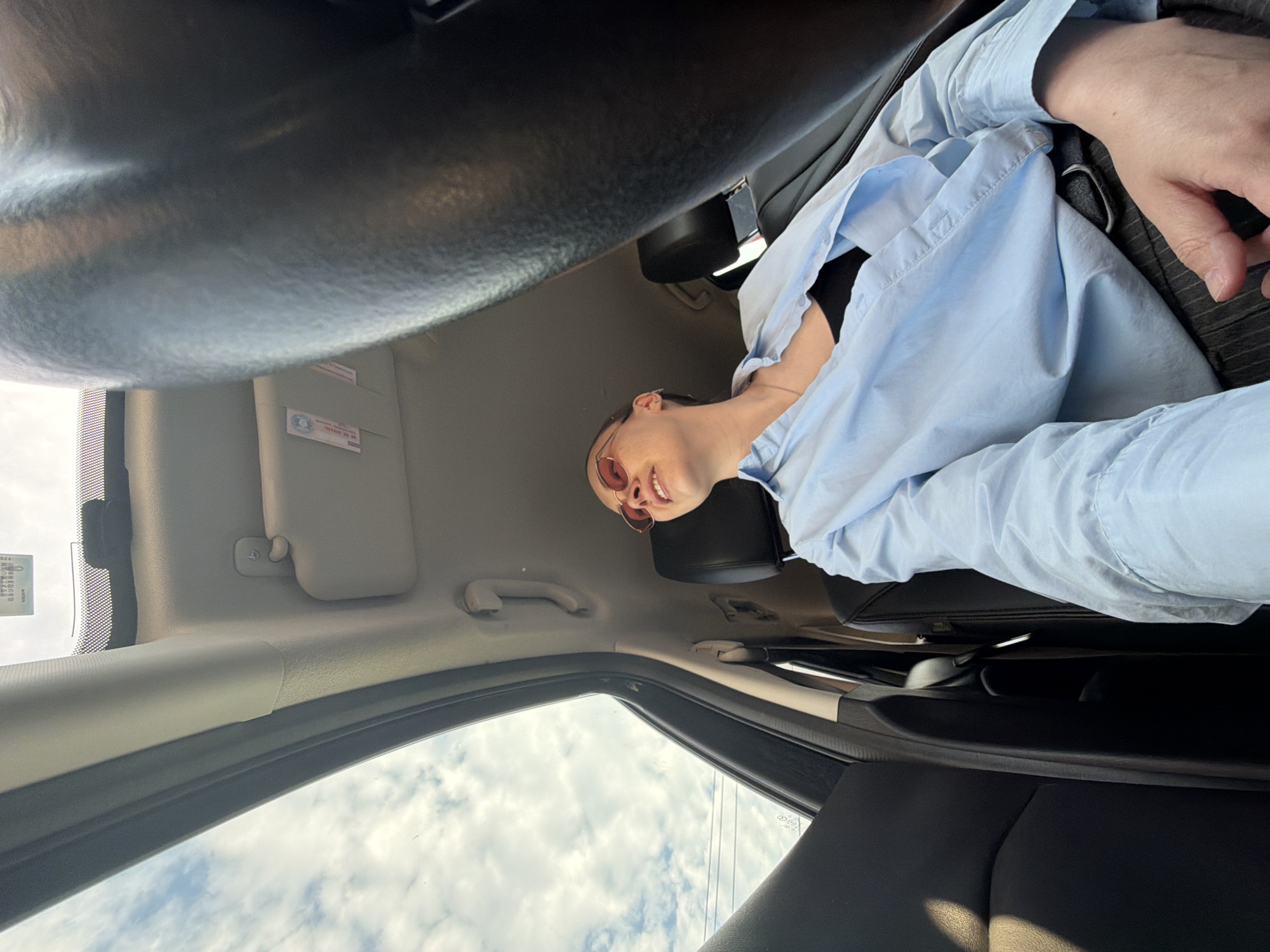The image shows the interior of a car, with a person seated in the driver's seat, wearing a light blue shirt and sunglasses. Part of the steering wheel is visible in the top left. The image is taken from an unusual angle, looking upwards towards the driver and the sky through the windshield. The location in Chaikovsky, Russia, cannot be determined from this image.