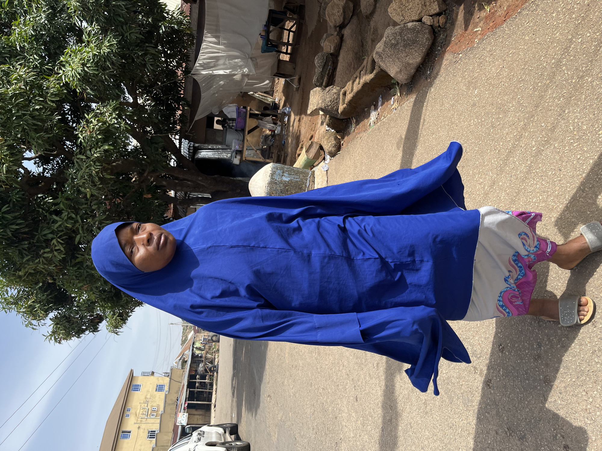A woman wearing a blue hijab and silver sandals stands on a paved road. Behind her is a tree on the left, and across the street is a two-story yellow building with a white car parked in front. In the background, there is a bustling market area with makeshift stalls and people. The scene appears to be in Jos, Nigeria, judging from the architectural style and overall environment.