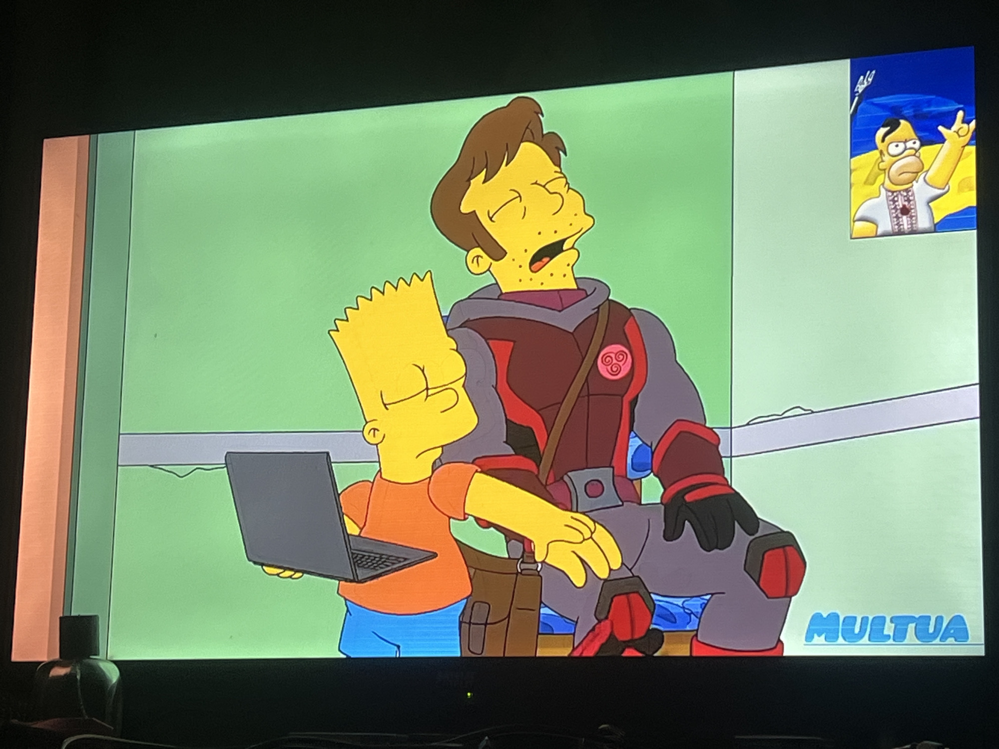 The image shows a scene from the animated television show "The Simpsons" playing on a television screen. In the foreground, Bart Simpson, dressed in an orange shirt and blue shorts, is holding a black laptop and appears to be interacting with another character. This character, who resembles a grown-up Bart Simpson, is wearing a futuristic-looking outfit in shades of gray and red, with a dark brown jacket and gloves. The background is a plain light green wall with a grey horizontal line running across it.
In the upper right corner of the screen, there is a smaller inset image of Homer Simpson wearing traditional Ukrainian attire, with the Ukrainian flag behind him. The logo "MULTUA" is visible in the bottom right corner of the main image. The image on the television is framed by the dark border of the television screen, with a portion of a speaker or stand visible at the bottom left. The overall scene is brightly lit, suggesting it is being viewed in a room with ambient light. The location mentioned in the prompt, Zoriane, Ukraine, is not depicted in the image itself, nor is there any visual information to suggest this specific geographical context.