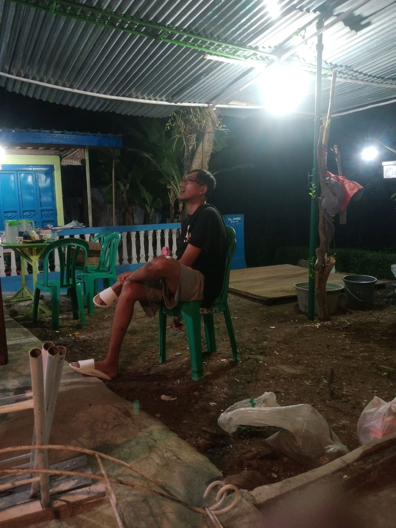 The image captures a man sitting on a green plastic chair outdoors at night in Rejasari, Indonesia. He is wearing glasses, a black t-shirt, and grey shorts. His left leg is crossed over his right, and he is wearing a white sandal on his right foot. The man is looking upwards and to his right.
The setting appears to be a covered outdoor area, possibly a terrace or an open-air dining space. The roof is made of corrugated metal and supported by metal beams painted green. Bright lights are suspended from the ceiling, illuminating the scene. Behind the man, there are more green plastic chairs and a green and yellow table with items on it, including a pitcher and what appears to be food containers. Further back, a building with blue doors and windows is visible, along with dense foliage.
The ground is dirt and gravel. Several objects are scattered around, including plastic bags, buckets, and what looks like a wooden platform. A pole with some clothing items hanging from it stands to the right of the man. In the foreground, some metal pipes and the edge of what might be a seating area or a railing are visible. The overall impression is a casual, somewhat cluttered outdoor gathering space at night.