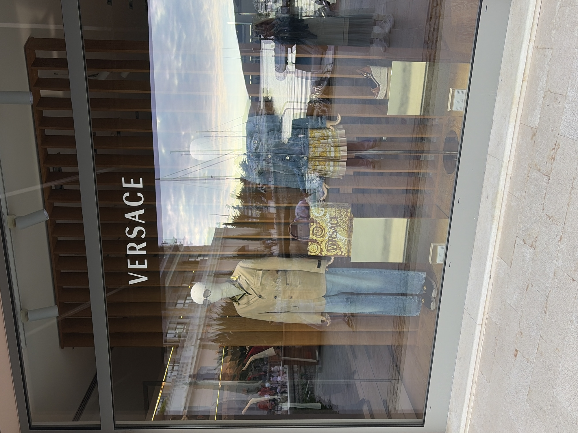 The image depicts the window display of a Versace store in Tivat, Montenegro. Mannequins are displayed with clothing and accessories reflecting the Versace brand. The store's facade is characterized by a modern design, incorporating horizontal wooden slats. Reflections in the glass display window reveal the outdoor environment, including people and boats, indicating the store's likely proximity to a marina or waterfront area. The surroundings suggest a well-maintained, upscale shopping area, consistent with the luxury brand's image.