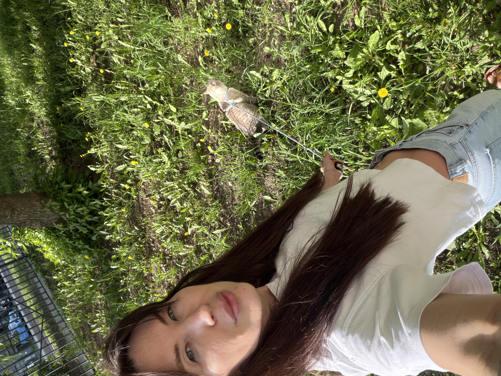 A selfie taken in Perm, Russia, shows a woman with long, dark hair and blue eyes looking at the camera. She is wearing a white t-shirt and blue jeans. In the background, there is a cat on a leash in a grassy area with dandelions. A fence and tree can be seen in the periphery.