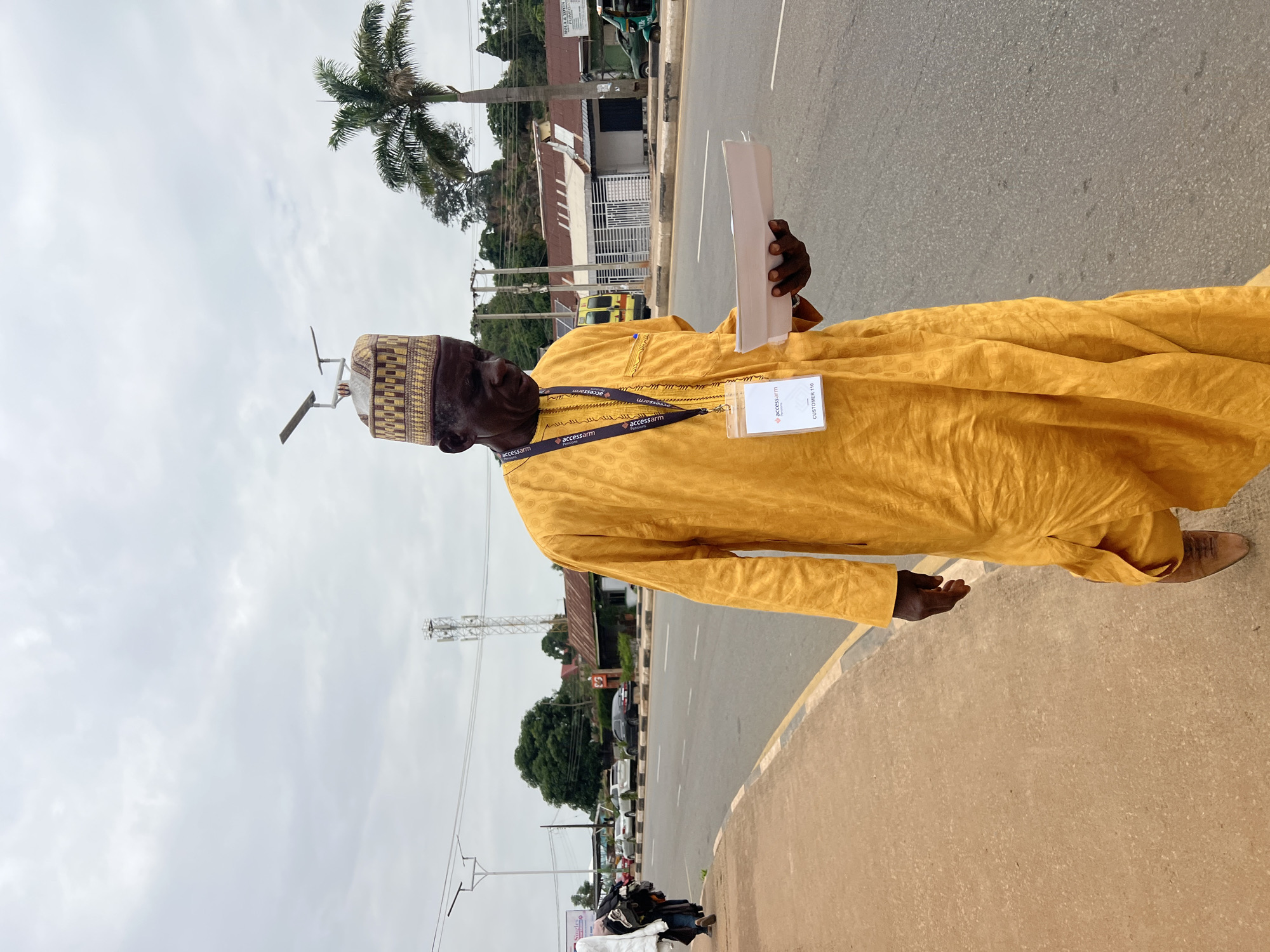 In Jos, Nigeria, an elderly man dressed in a bright yellow traditional outfit and a patterned cap walks on the sidewalk next to a road. He holds a light-colored shallow dish in his hands. A lanyard with an ID badge labeled "Access Arm" and "Customer 110" hangs around his neck. The road is paved and has a yellow and black border. Cars are visible on the road, and buildings with red roofs and various signs line the opposite side. A tall palm tree stands in the background, and the sky is overcast. The scene appears to be a street in a moderately busy urban area.