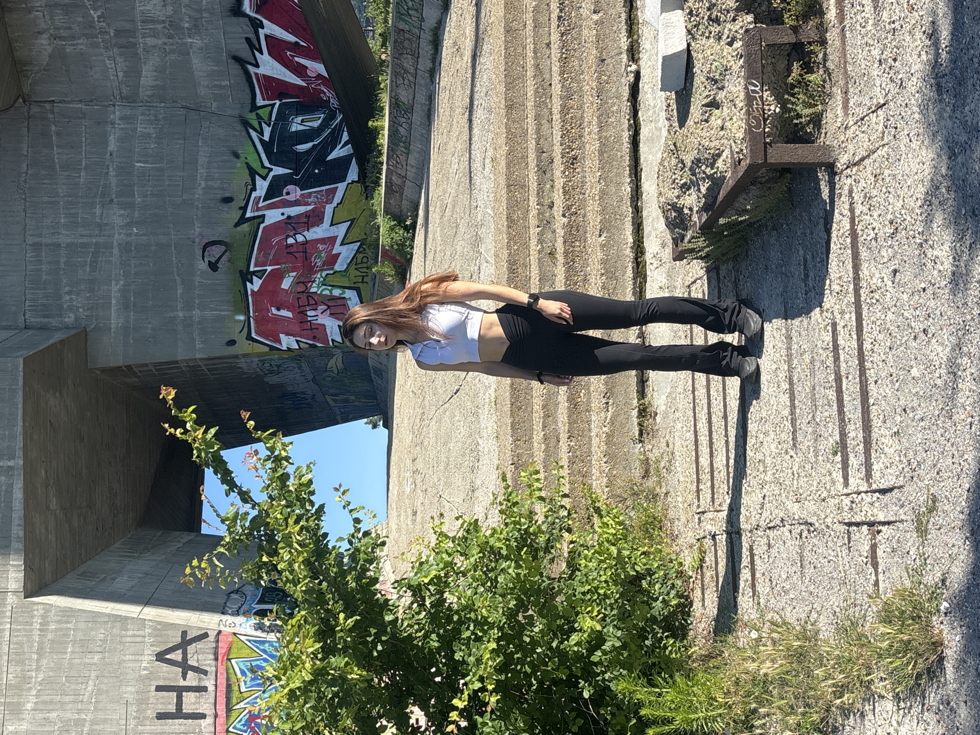 A young woman with long brown hair stands on a concrete staircase outdoors. She is wearing a white crop top and black leggings. The background features a large concrete structure with graffiti. The location is under the Asparuhov Bridge in Varna, Bulgaria. The area appears to be under construction or abandoned, with overgrown vegetation and debris scattered around. The lighting suggests it is daytime, with clear sunlight casting shadows.