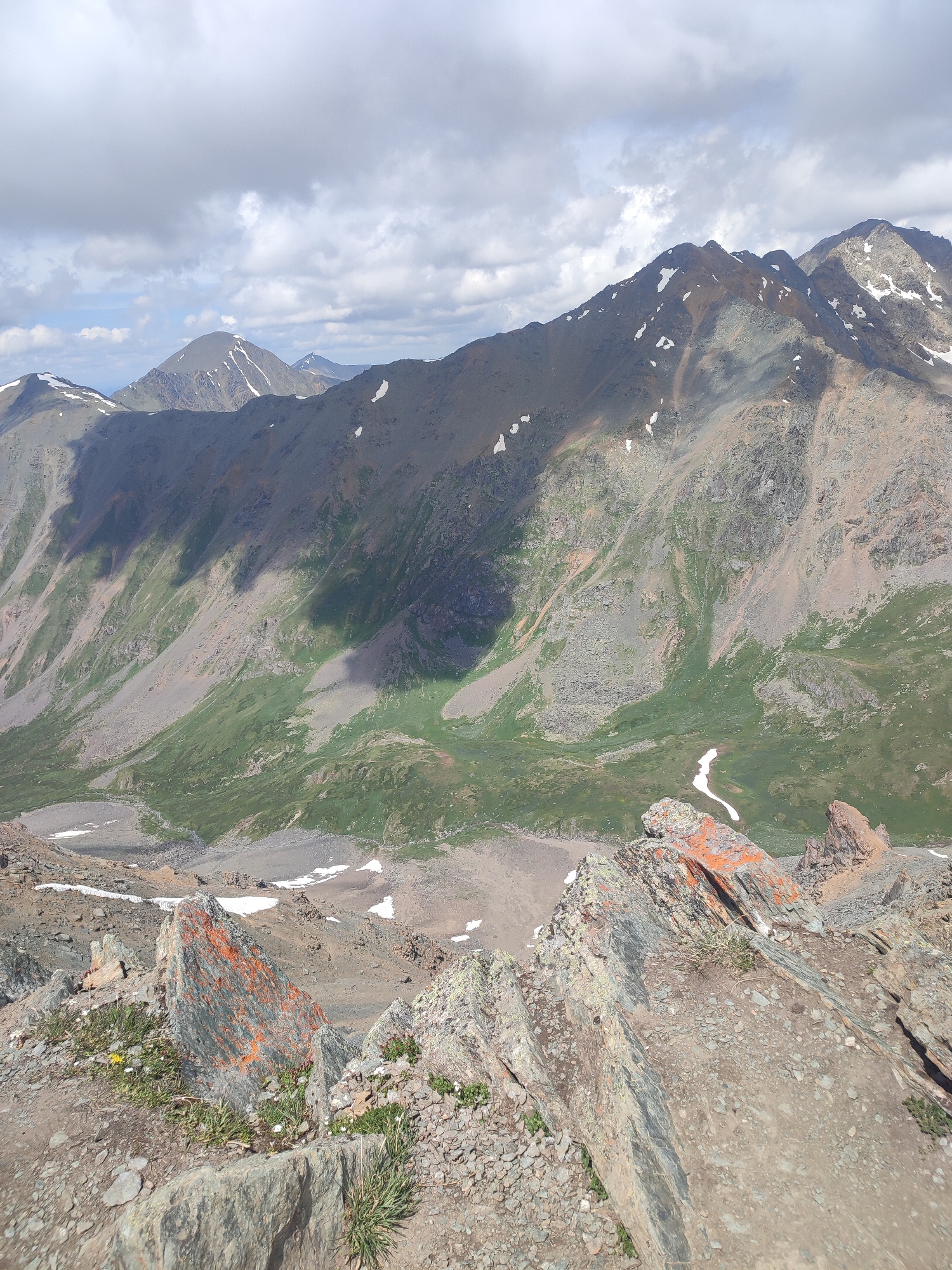 This is an aerial, eye-level, full outdoor shot of a mountainous landscape under a partly cloudy sky. The foreground consists of rocky terrain with sparse green vegetation and some larger, reddish-orange rocks. Beyond the foreground, the scene opens up to a vast mountain valley with green slopes interspersed with rocky scree and patches of snow. In the background, several mountain peaks rise, some capped with snow, under a dramatic sky with heavy clouds. The overall impression is one of a rugged, natural environment, likely at a high altitude. The location is identified as Aktash, Russia. There are no people or animals visible in the image, nor any man-made structures or objects.