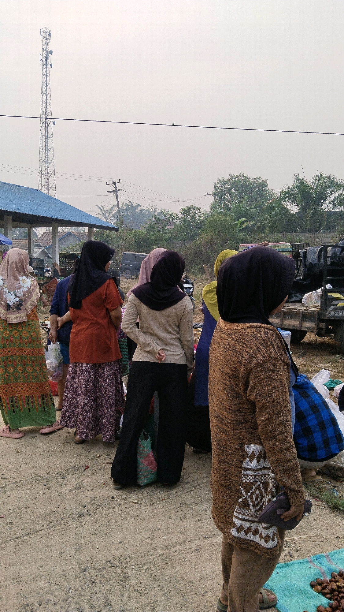 In Marga Sakti, Indonesia, a group of women are gathered outdoors, facing away from the camera. The scene appears to be a market or gathering area, with a dirt ground and a few structures in the background. A tall cell tower dominates the sky, with a bird perched on a wire in the foreground. The women are wearing traditional Islamic clothing, including hijabs and long dresses or skirts. Some are carrying bags, and one is holding a small purse. The overall atmosphere is casual and everyday.