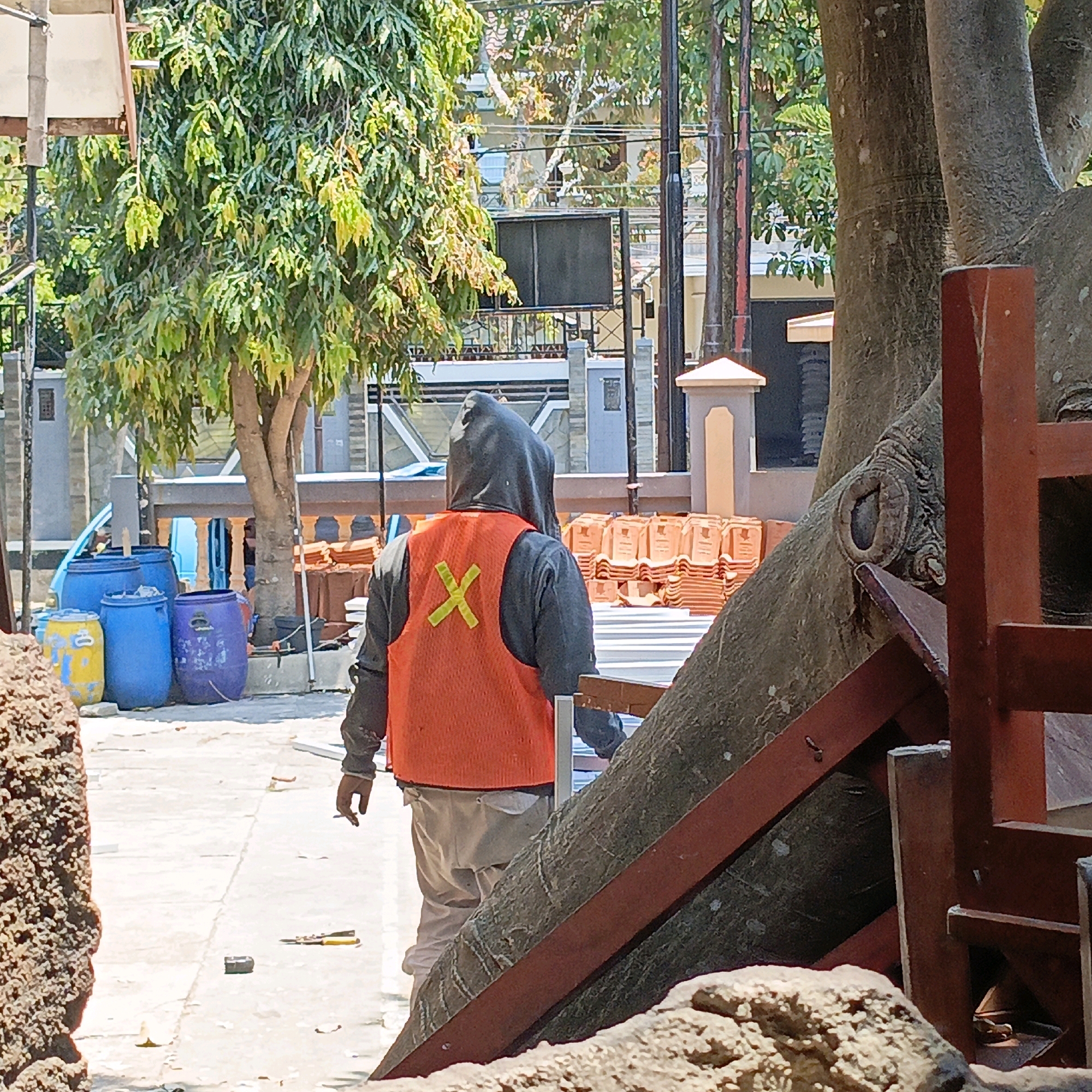 The image depicts a sunny outdoor scene in Salatiga, Indonesia, likely a construction or work area. In the foreground, a person wearing a grey hooded sweatshirt and an orange reflective vest with a yellow "X" marking on the back is walking away from the camera. They are also wearing light-colored pants and appear to be smoking a cigarette. To the left of the person, there are several large blue plastic barrels and one yellow and blue barrel. In the background, there is a large tree with a thick trunk, stacks of orange roof tiles, and various structures, including fences and buildings. The ground is paved and appears to be a walkway or road. The overall impression is one of ongoing work or activity in a somewhat cluttered outdoor environment.