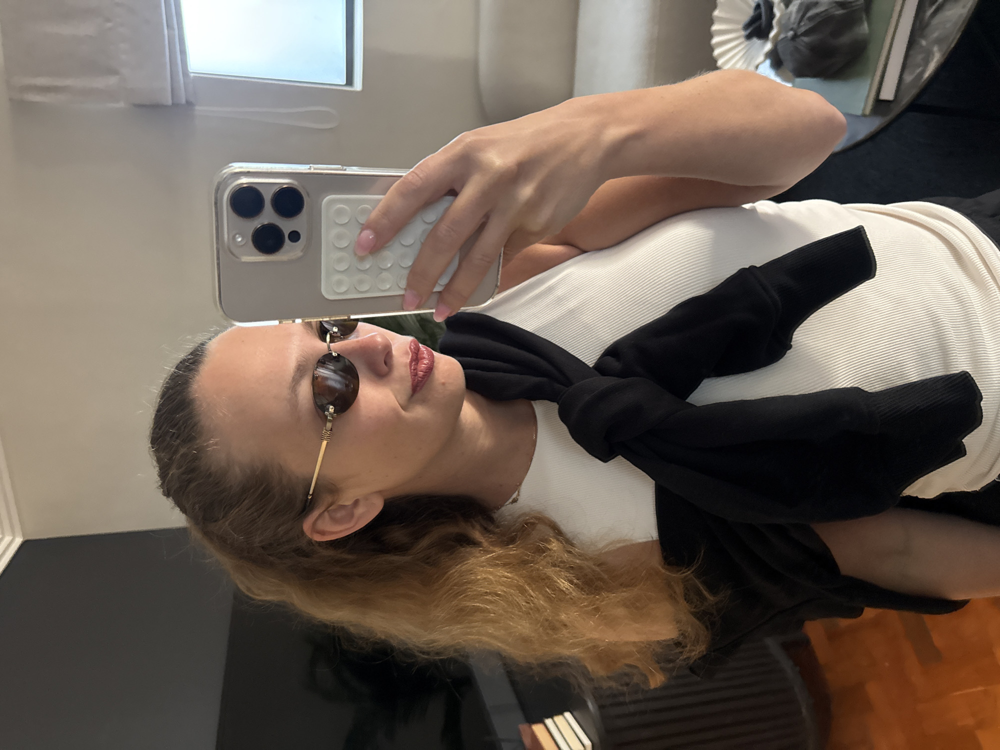 A young woman is captured taking a mirror selfie indoors. She has light brown or blonde, wavy hair and is wearing stylish dark sunglasses with slender gold frames. Her lips are painted with a reddish-pink lipstick, and her expression is relaxed as she gazes towards her reflection. She is dressed in a white, ribbed tank top or camisole, with a black long-sleeved garment, possibly a sweater or jacket, tied casually around her chest.
In her right hand, she holds a silver or light-colored smartphone, likely an iPhone due to its triple-lens camera array, encased in a clear protective cover. A white accessory, resembling a grip or multi-point PopSocket, is attached to the back of the phone. Her left hand is also visible near the phone, positioned to support her hold.
The background shows an interior setting, possibly a room in a home or hotel. A window with light-colored blinds or a sheer curtain is visible, allowing natural light to illuminate the space, suggesting it is daytime. Blurry elements in the background include light-colored furniture, perhaps a sofa or bed, and darker surfaces like a table. The overall ambiance is casual and personal. Although not visually depicted, the image is situated in Cape Town, South Africa. There is no discernible text visible in the frame.