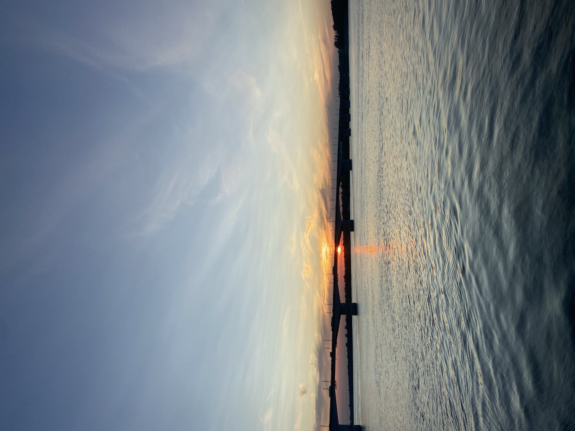 The image shows a wide river with a bridge crossing it. The sun is setting, casting a warm glow on the sky and reflecting on the water. There are some clouds in the sky, streaked with orange and pink hues. The bridge is long and made of concrete, with several pillars supporting it. Cars can be seen driving on the bridge. The overall atmosphere is peaceful and serene. This scene is likely the Kama River in the city of Perm, Russia, given the prominent bridge and wide river, characteristics of the city's geography.