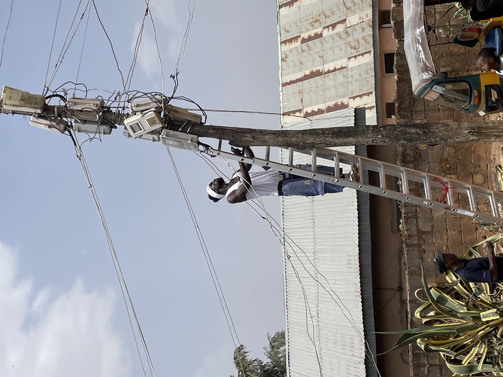 An individual is working on a utility pole with a ladder in Jos, Nigeria. The pole supports numerous electrical meters and wires. The worker is wearing a white shirt, jeans, and a head covering. A corrugated metal structure stands in the background. Nearby, there is a person sitting in a green and yellow motorized vehicle and another person standing next to an agave plant.