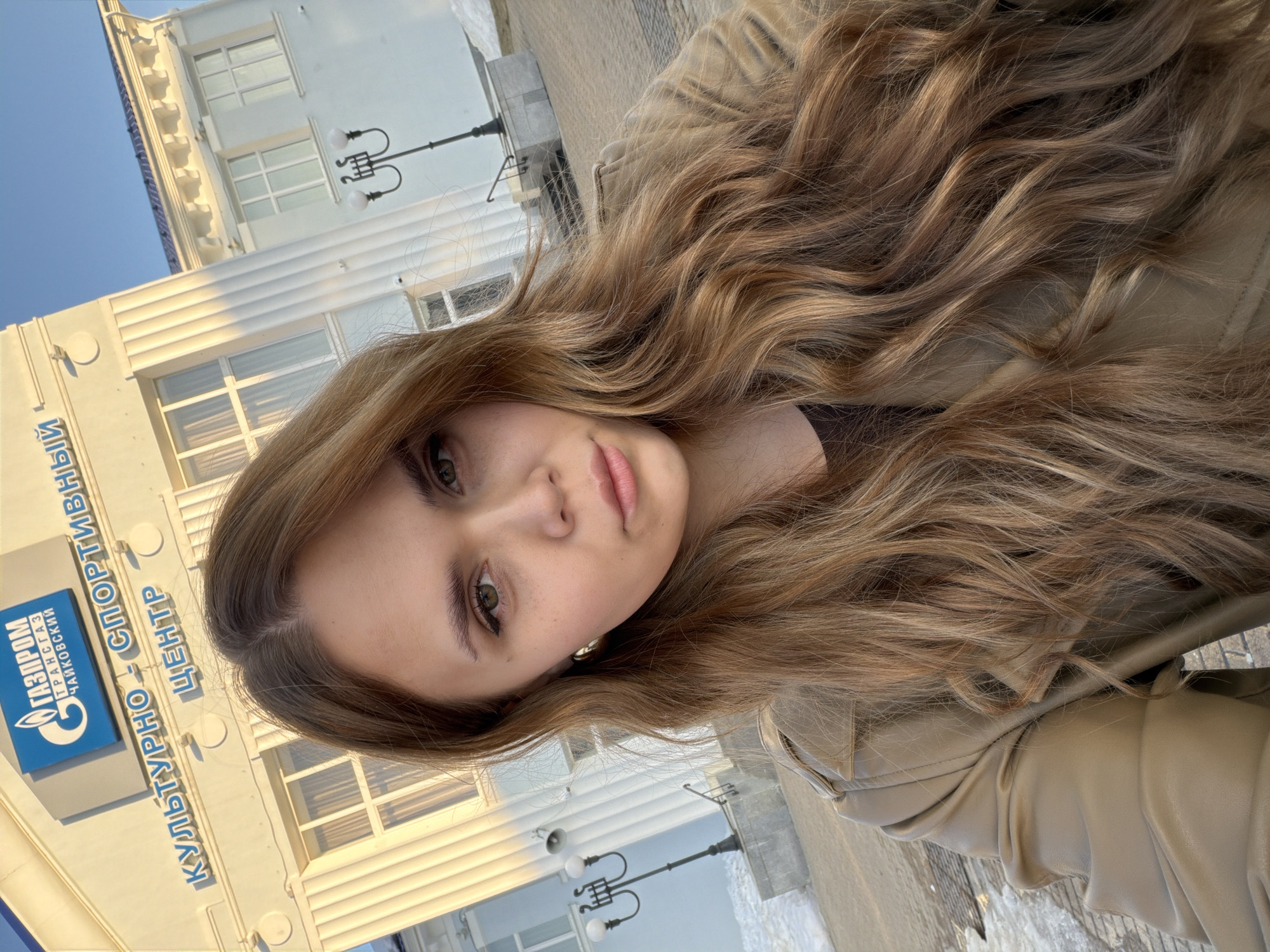 A selfie of a young woman, likely in her late teens or early twenties, with long, wavy, light brown and blonde hair. She has fair skin and green eyes. She wears a light tan jacket. She is standing outdoors, likely on a sidewalk. Behind her is a building with a sign in Russian that includes the words "ГАЗПРОМ" (Gazprom) and "СПОРТИВНЫЙ ЦЕНТР" (Sports Center). Based on the text of the sign that says "Газпром трансгаз Чайковский", the location is in the city of Chaikovsky, Perm Krai, Russia. The building is light-colored, has multiple windows, and is accented with decorative features like columns and street lamps. The sky is blue, and it appears to be a clear day.