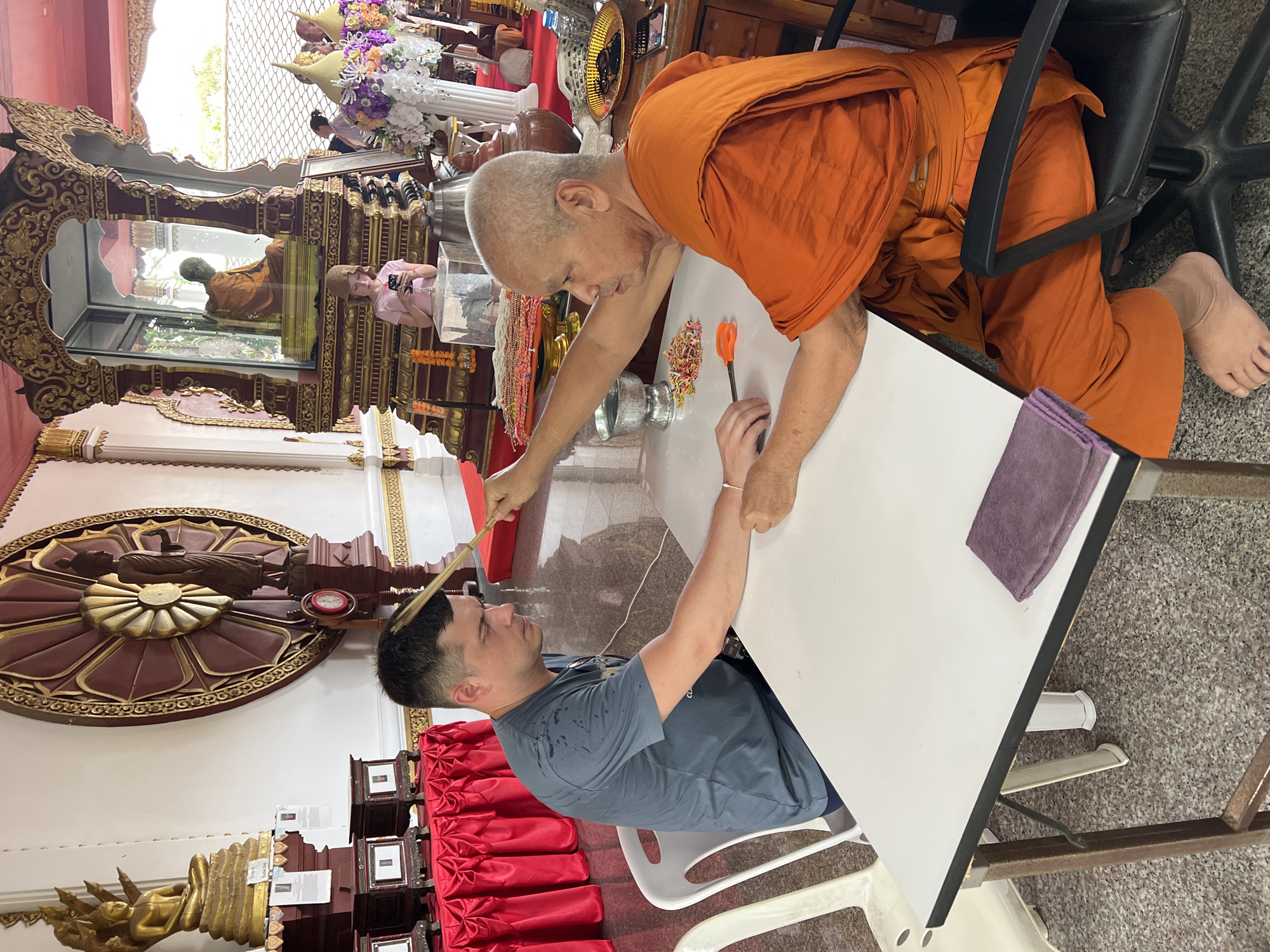 In Baan Hua Thanon, Thailand, within a temple setting, a man in a grey t-shirt sits at a white table facing a Buddhist monk in orange robes. The monk, with a shaved head, is holding a bundle of incense sticks and appears to be performing a ritual, possibly a blessing or a ceremony, on the man's head. The man is looking down with his eyes closed, his right arm resting on the table. In front of the monk are a pair of orange scissors and a small pile of colorful beads or flower petals. The table is white with a metal frame, and a purple cloth is placed on it. In the background, a large wooden wheel with intricate carvings is visible on the wall, and statues of Buddha are displayed on red-covered pedestals. A young girl is also visible in the background, looking at her phone. The environment suggests a sacred and traditional place of worship.