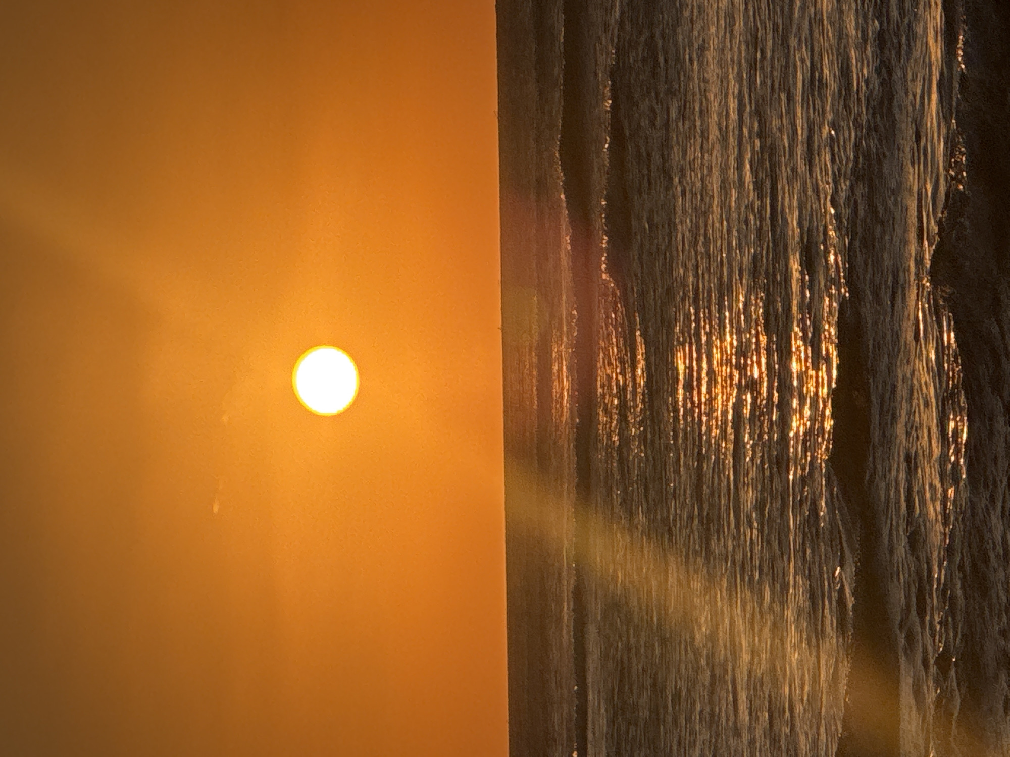 The image, taken in Kuta Selatan, Indonesia, depicts a vibrant sunrise or sunset over the ocean, with the scene oriented such that the sun is on the left and the water on the right (if viewed horizontally). A bright, circular sun, radiating intense golden-orange light, is prominently featured on the left side, casting a noticeable lens flare that stretches across the frame. The sky around the sun is painted in warm hues of orange and yellow, indicating either dawn or dusk.
On the right side, the surface of the water, likely the ocean, is illuminated by the sun's low angle. The water features numerous shimmering vertical reflections, appearing as golden streaks against the darker water, suggesting gentle waves or ripples catching the sunlight. These reflections create a textured, almost glowing effect on the water's surface. The overall color palette is dominated by warm oranges, yellows, and the contrasting darker tones of the water. No individuals or man-made objects are visible, and no text is present in the scene. The image captures a serene natural moment during golden hour in a coastal setting.