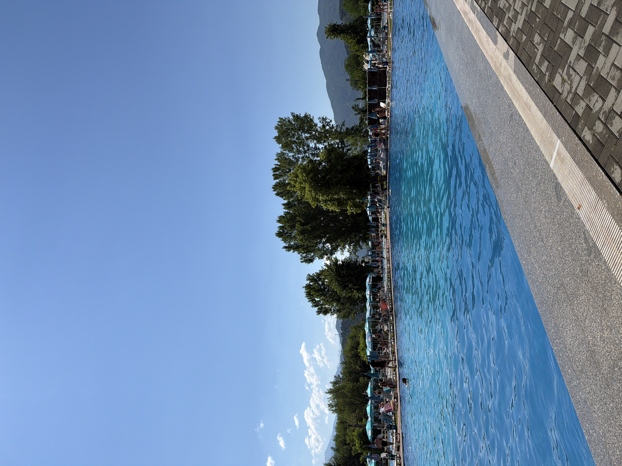 This is a wide, outdoor, eye-level shot of a swimming pool area on a clear, sunny day. The pool is a vibrant blue, with rippling water. In the foreground, the edge of the pool is a granite-like material, bordered by a paved walkway with a herringbone pattern. Beyond the pool, rows of lounge chairs are set up, many covered by green umbrellas. People are scattered throughout the lounging area, some sitting, some standing, and a few are in the pool. Behind the lounge chairs and umbrellas, a line of green trees forms a backdrop, with mountains visible in the distance under a clear blue sky. The location is identified as Simitli, Bulgaria.