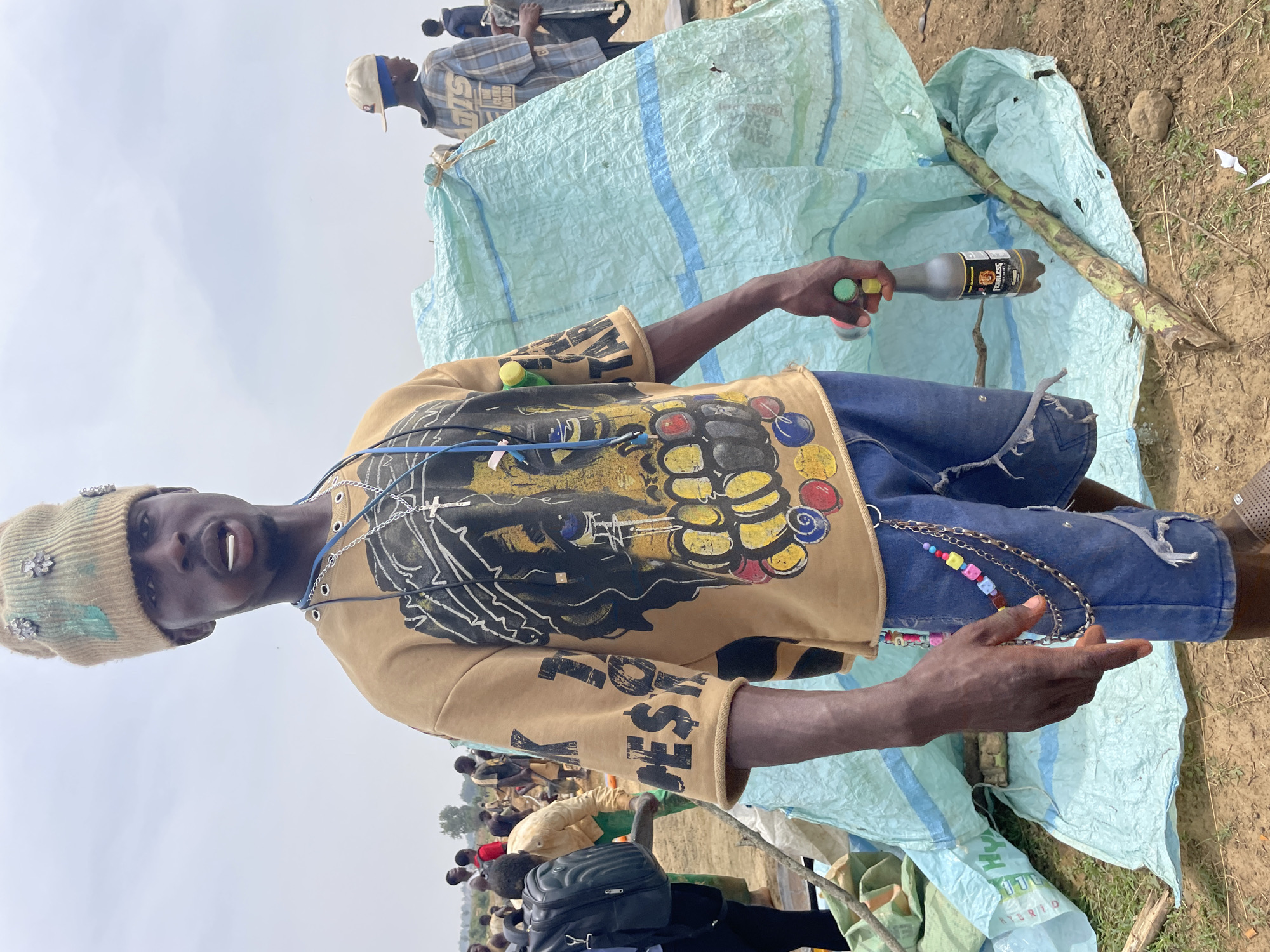 A man stands in a field, holding a bottle of "Fearless" drink in his left hand. He is wearing a decorated knit cap, a tan long-sleeved shirt with an image of a lion and the word "Fearless" on the sleeve, and blue denim shorts. A silver chain with colorful beads is attached to his shorts. Behind him is a pile of material covered with a light blue tarp with blue stripes. In the background, other people are visible. The location is Maiyanga, Nigeria. The ground is dirt and grass.
