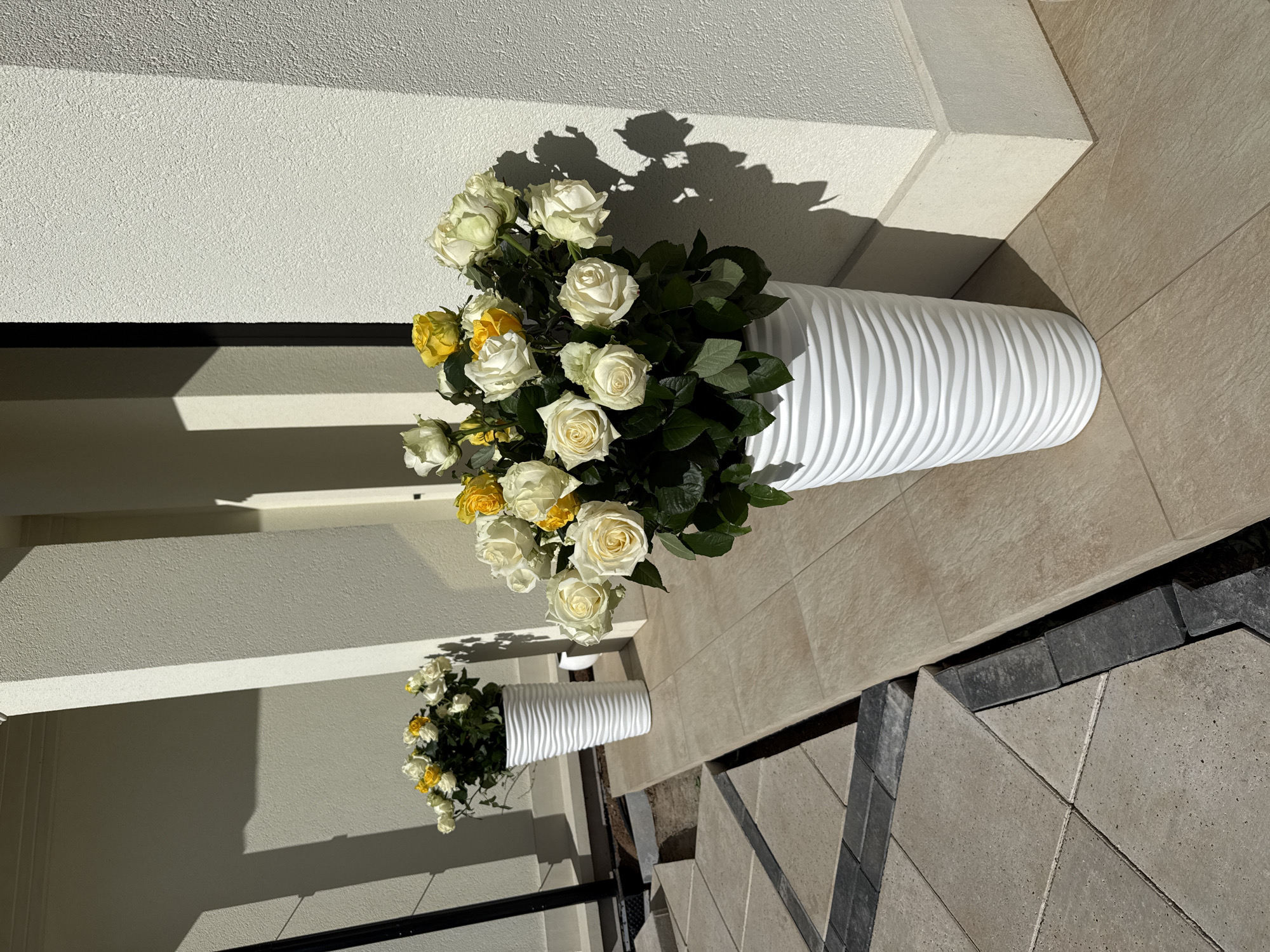 The image shows a high-angle view of two white vases filled with white and yellow roses, positioned outside a building. The vases are cylindrical with a textured, wavy surface. The larger vase is at the top of the frame, casting a shadow on the wall behind it. The smaller vase is located towards the lower left corner. The background consists of a light-colored tiled floor and a white wall. A black railing is visible in the lower left corner. The scene appears to be well-lit, with the shadows indicating a sunny day. There is no evidence that this picture was taken in the city of Гольяны, Russia.
