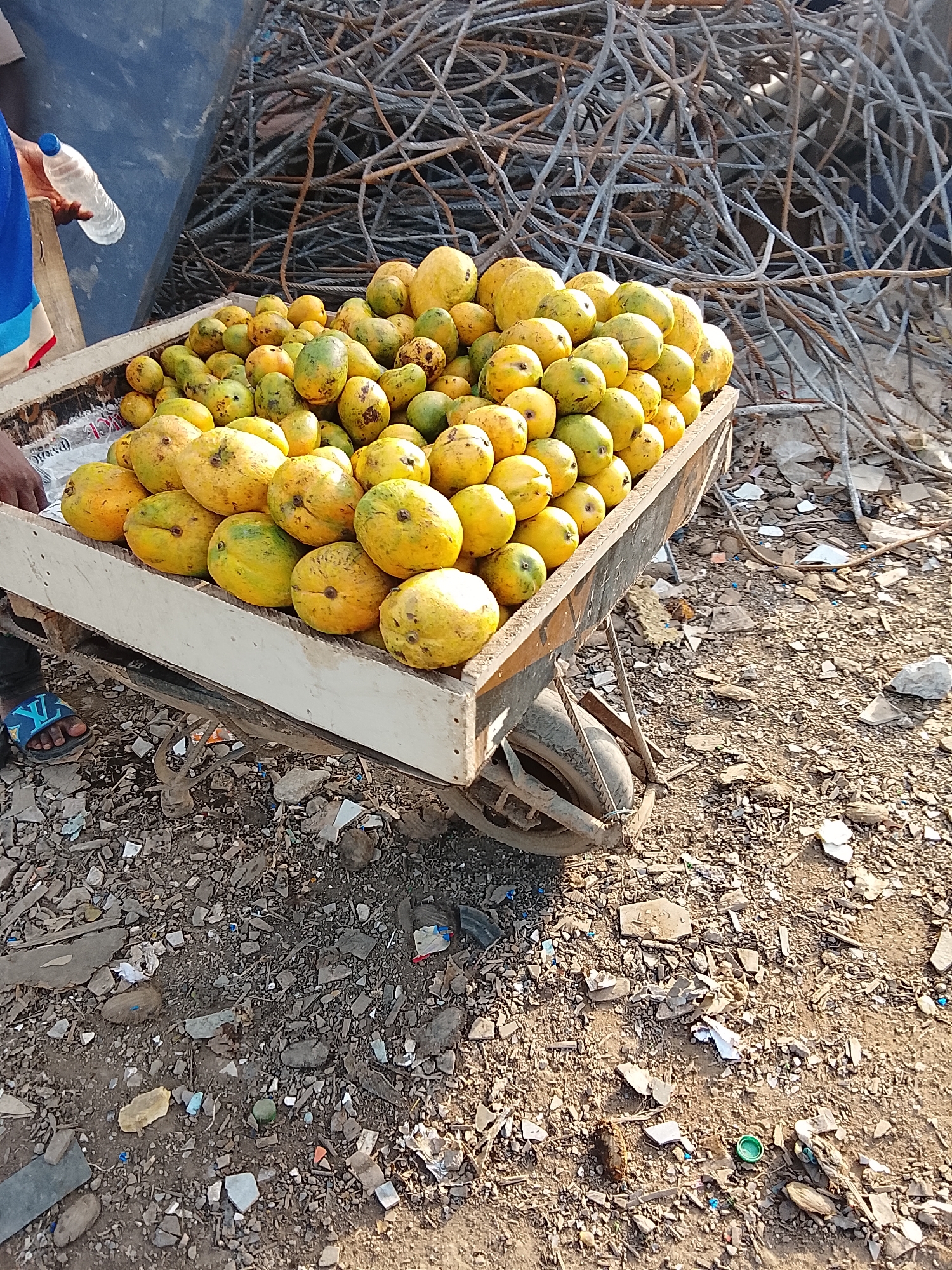 The image shows a cart laden with mangoes in an apparent outdoor marketplace or roadside setting in Abuja, Nigeria. The cart, with white wooden sides, is piled high with ripe mangoes. Adjacent to the cart is a large pile of rusted metal rods, possibly reinforcing bars. The ground is covered in dirt, small rocks, and scattered debris, suggesting an informal or undeveloped area. A person wearing a blue shirt and holding a water bottle stands next to the cart. Part of their foot, clad in a blue sandal, is visible.